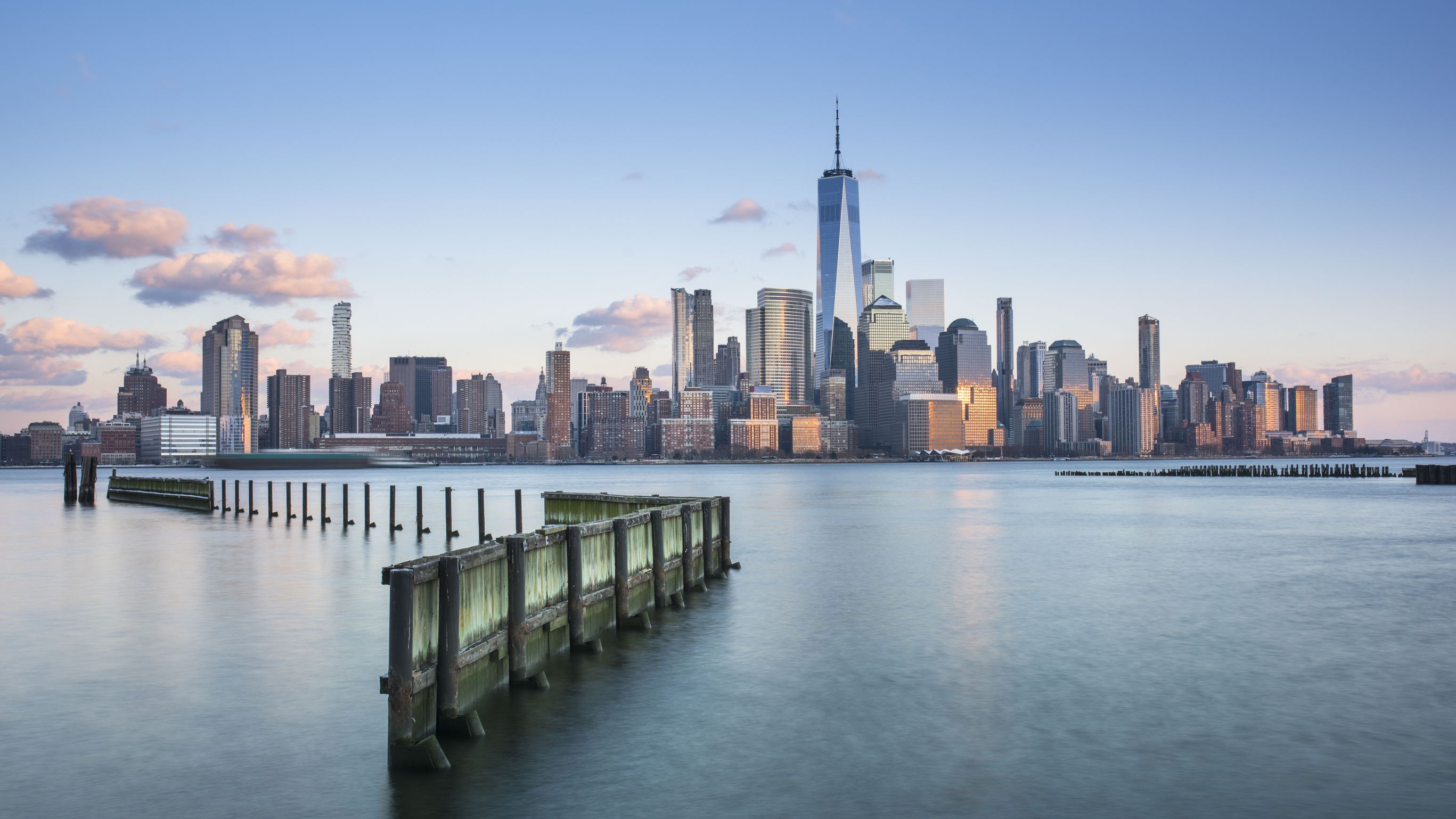Blick auf die Skyline von Manhattan bei klarem Himmel, mit dem markanten One World Trade Center und ruhigem Wasser im Vordergrund, das eine alte Holzpier umspült. Blick auf die Skyline von Manhattan bei klarem Himmel, mit dem markanten One World Trade Center und ruhigem Wasser im Vordergrund, das eine alte Holzpier umspült.