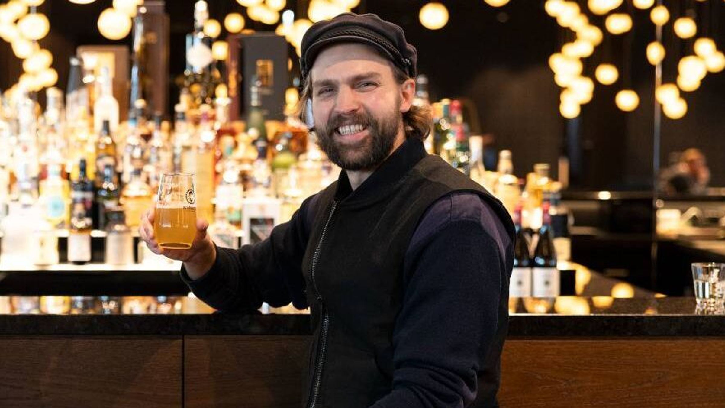 Mann in legerer Kleidung sitzt an der Bar und hält lächelnd ein Glas Bier, warme Beleuchtung im Hintergrund schafft ein einladendes Ambiente.