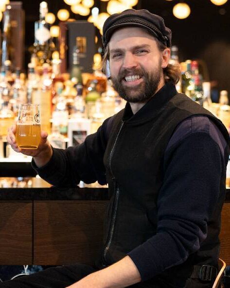 Mann in legerer Kleidung sitzt an der Bar und hält lächelnd ein Glas Bier, warme Beleuchtung im Hintergrund schafft ein einladendes Ambiente.