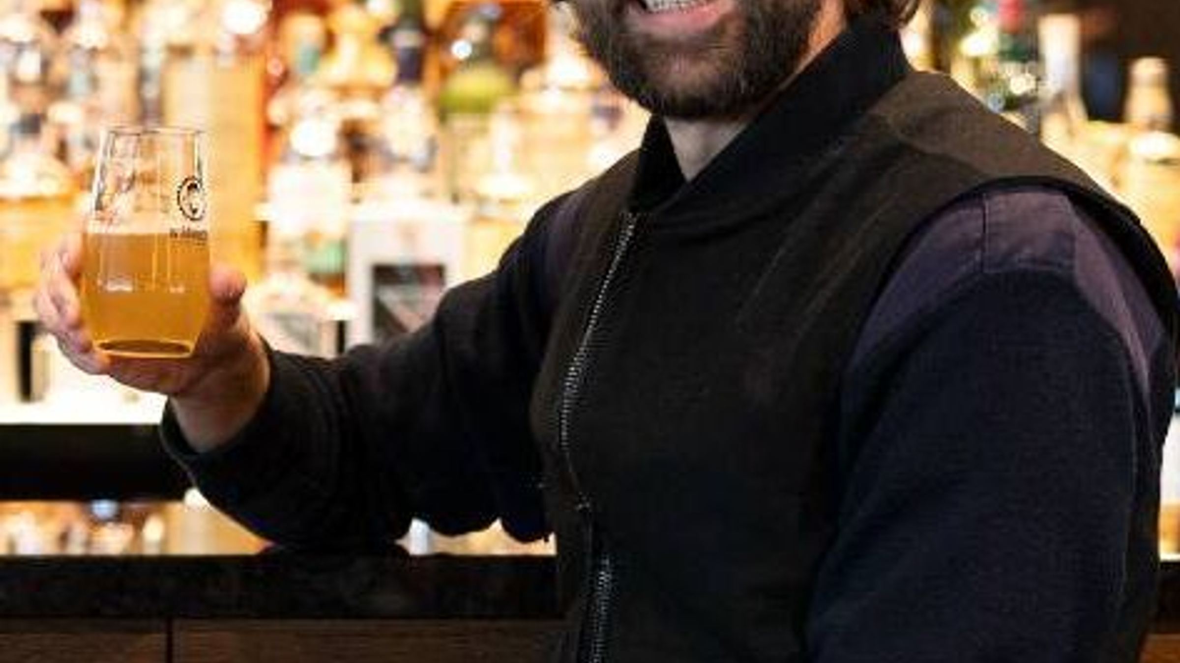 Mann in legerer Kleidung sitzt an der Bar und hält lächelnd ein Glas Bier, warme Beleuchtung im Hintergrund schafft ein einladendes Ambiente. Mann in legerer Kleidung sitzt an der Bar und hält lächelnd ein Glas Bier, warme Beleuchtung im Hintergrund schafft ein einladendes Ambiente.