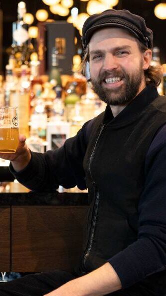 Mann in legerer Kleidung sitzt an der Bar und hält lächelnd ein Glas Bier, warme Beleuchtung im Hintergrund schafft ein einladendes Ambiente.