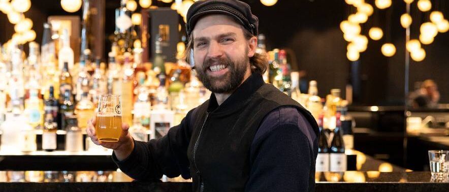 Mann in legerer Kleidung sitzt an der Bar und hält lächelnd ein Glas Bier, warme Beleuchtung im Hintergrund schafft ein einladendes Ambiente.
