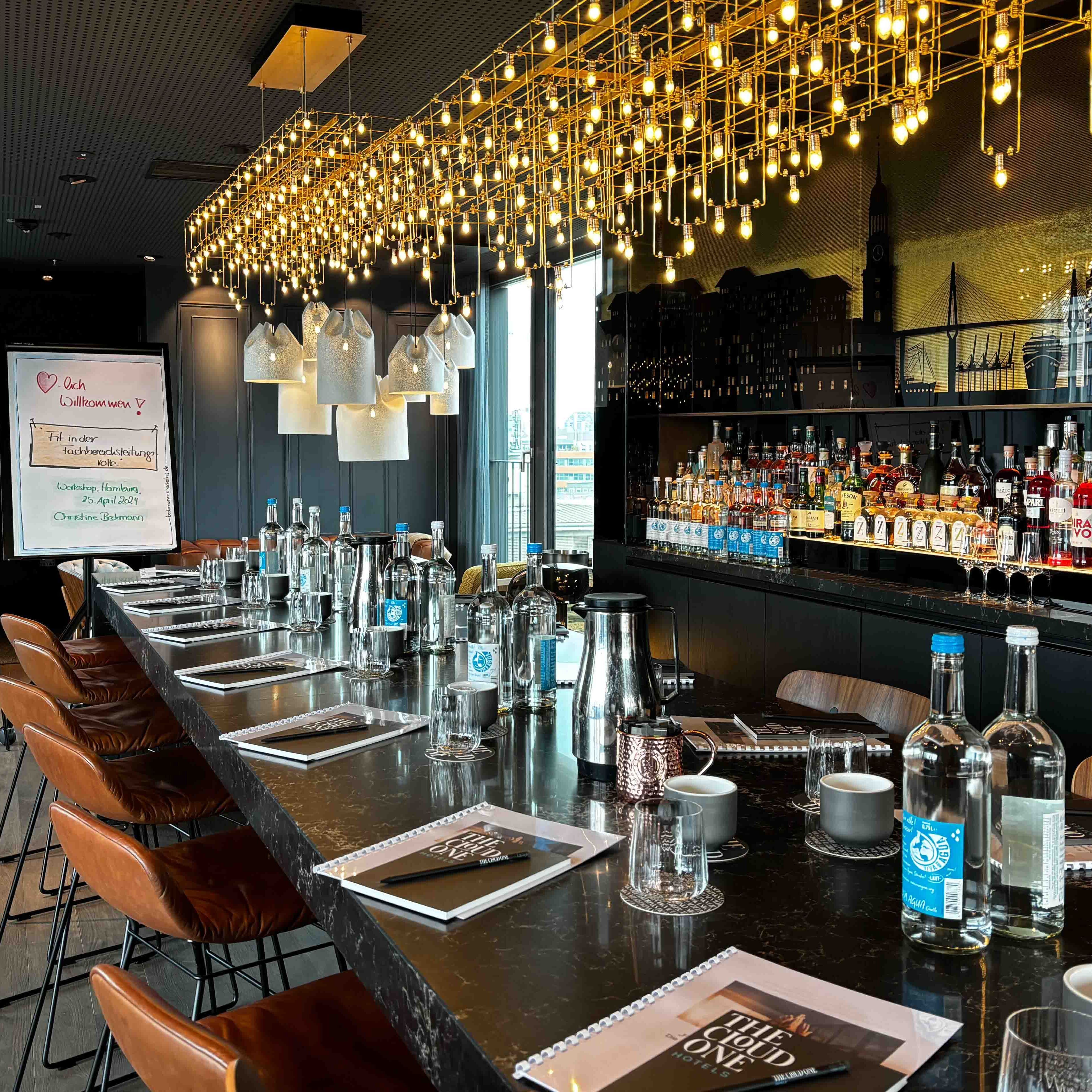 Motel One Hotelbar mit langer Theke, bequemen Lederhockern, stilvoller Beleuchtung und umfangreicher Auswahl an Spirituosen, einladend für gesellige Abende oder entspannte Drinks.
