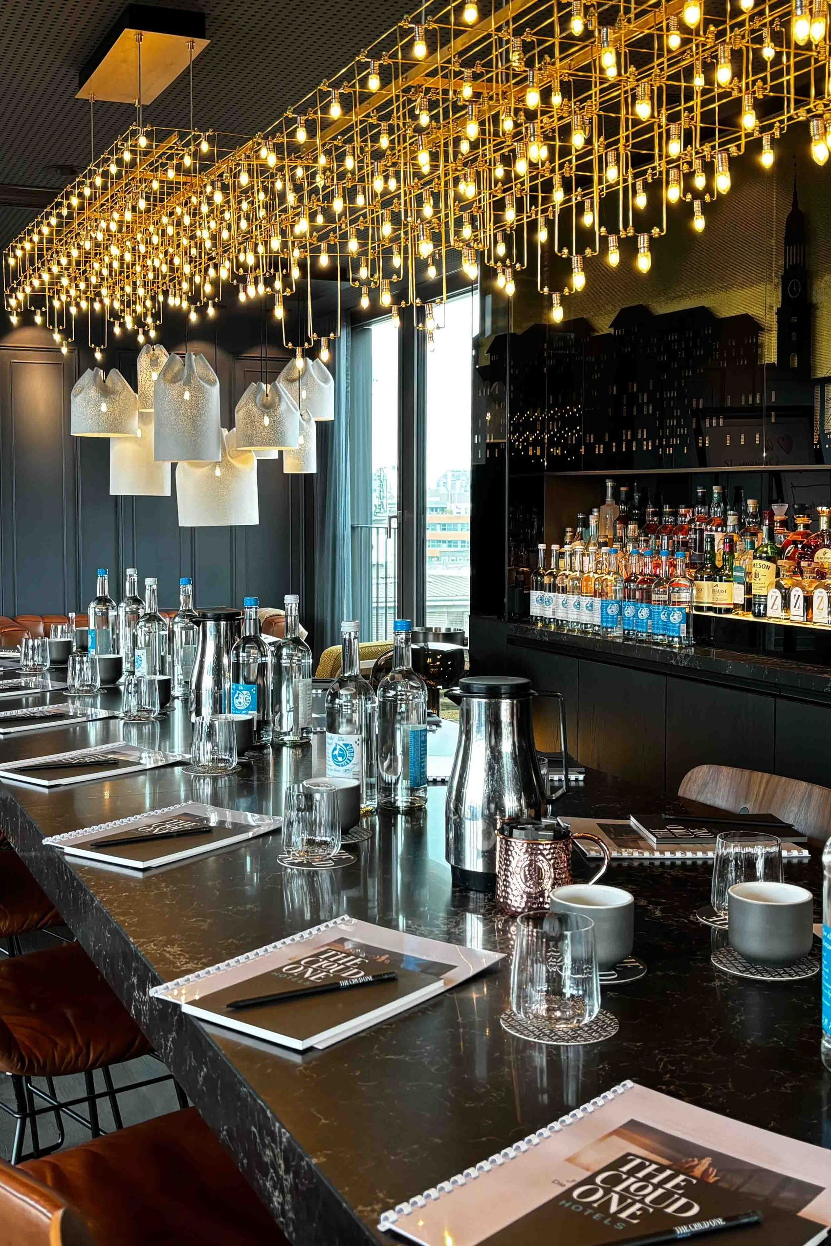 Motel One Hotelbar mit langer Theke, bequemen Lederhockern, stilvoller Beleuchtung und umfangreicher Auswahl an Spirituosen, einladend für gesellige Abende oder entspannte Drinks.