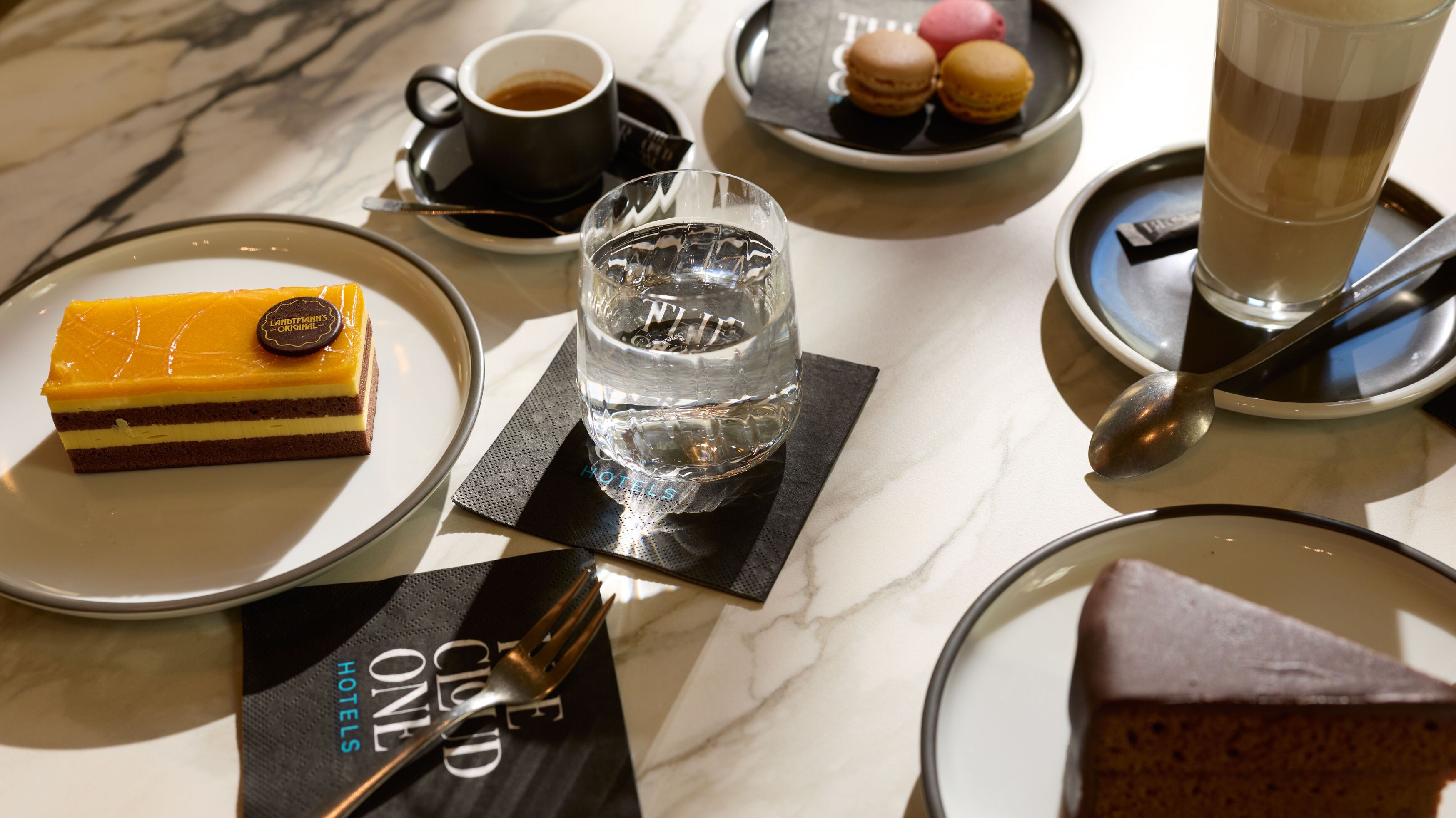 Verschiedene süße Köstlichkeiten auf Tellern, darunter ein Schokoladenkuchen, ein Schichtdessert mit gelber Glasur und bunt gefärbte Macarons, begleitet von einer Tasse Kaffee, einem Glas Wasser und einem Latte Macchiato auf einem Marmor-Tisch.