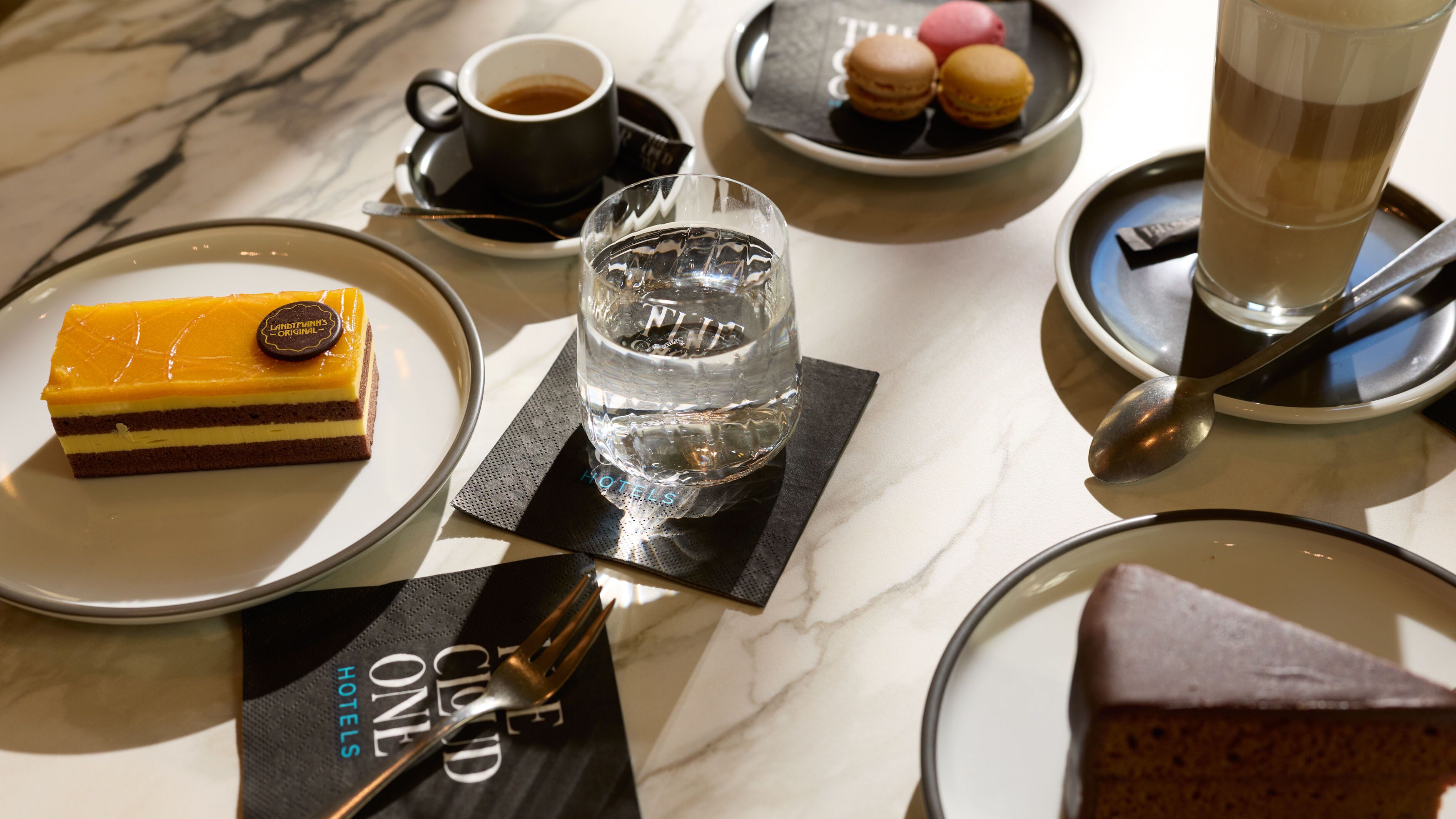 Verschiedene süße Köstlichkeiten auf Tellern, darunter ein Schokoladenkuchen, ein Schichtdessert mit gelber Glasur und bunt gefärbte Macarons, begleitet von einer Tasse Kaffee, einem Glas Wasser und einem Latte Macchiato auf einem Marmor-Tisch.