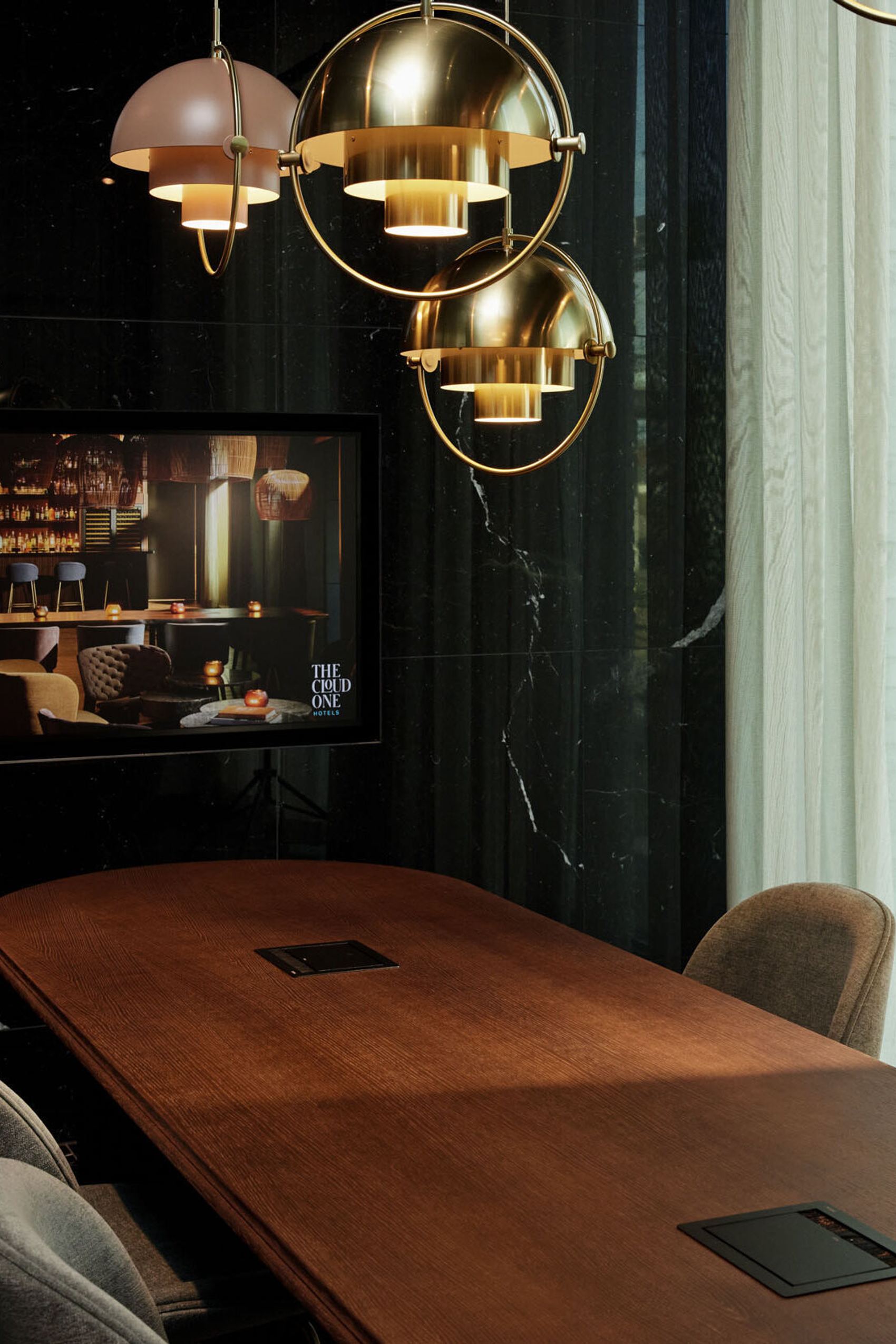 Motel One Konferenzraum mit langem Holztisch, bequemen Stühlen und großen Fenstern, die Tageslicht hereinlassen. Stilvolle goldene Hängeleuchten pendeln von der Decke.