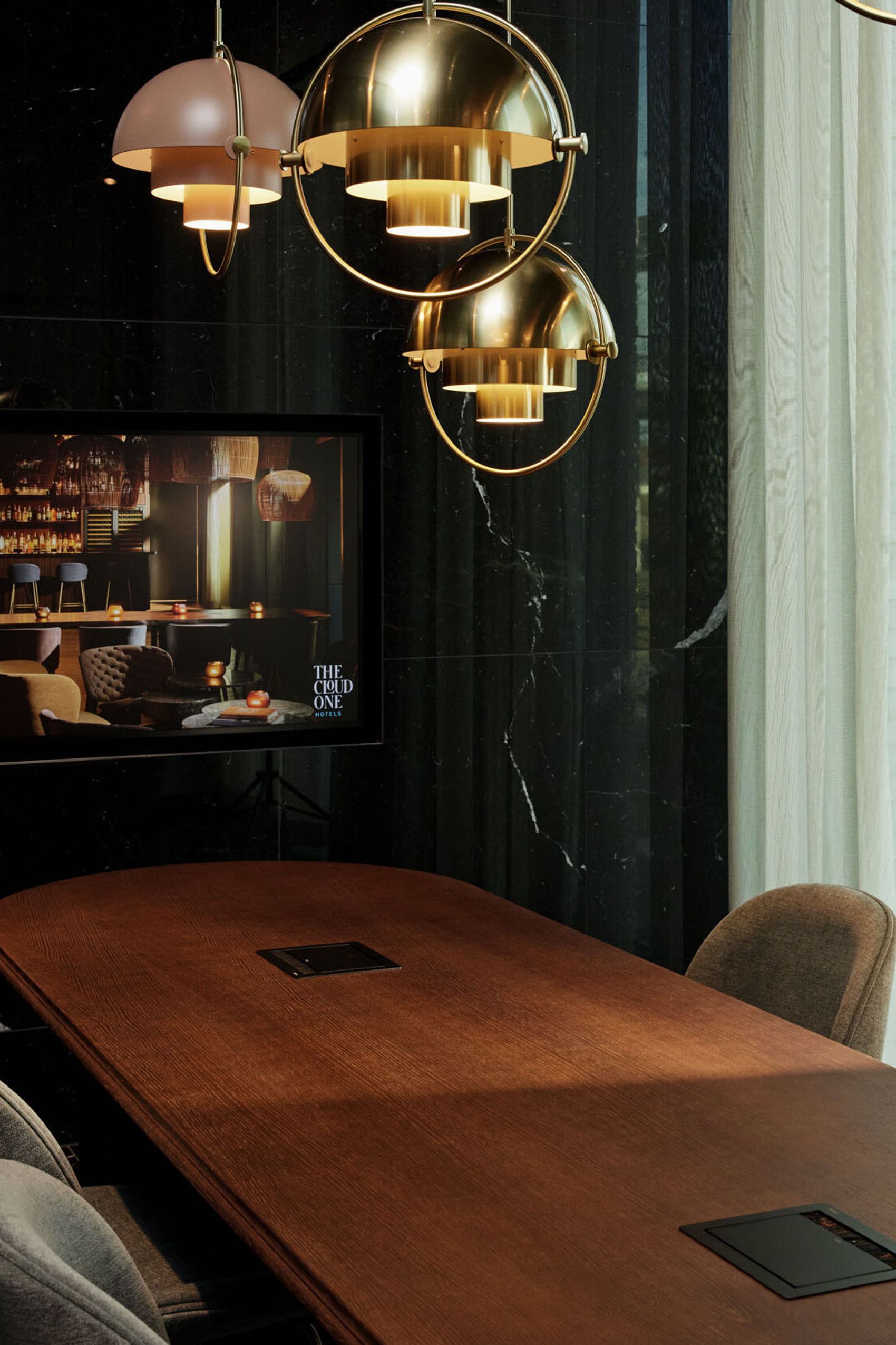 Motel One Konferenzraum mit langem Holztisch, bequemen Stühlen und großen Fenstern, die Tageslicht hereinlassen. Stilvolle goldene Hängeleuchten pendeln von der Decke.