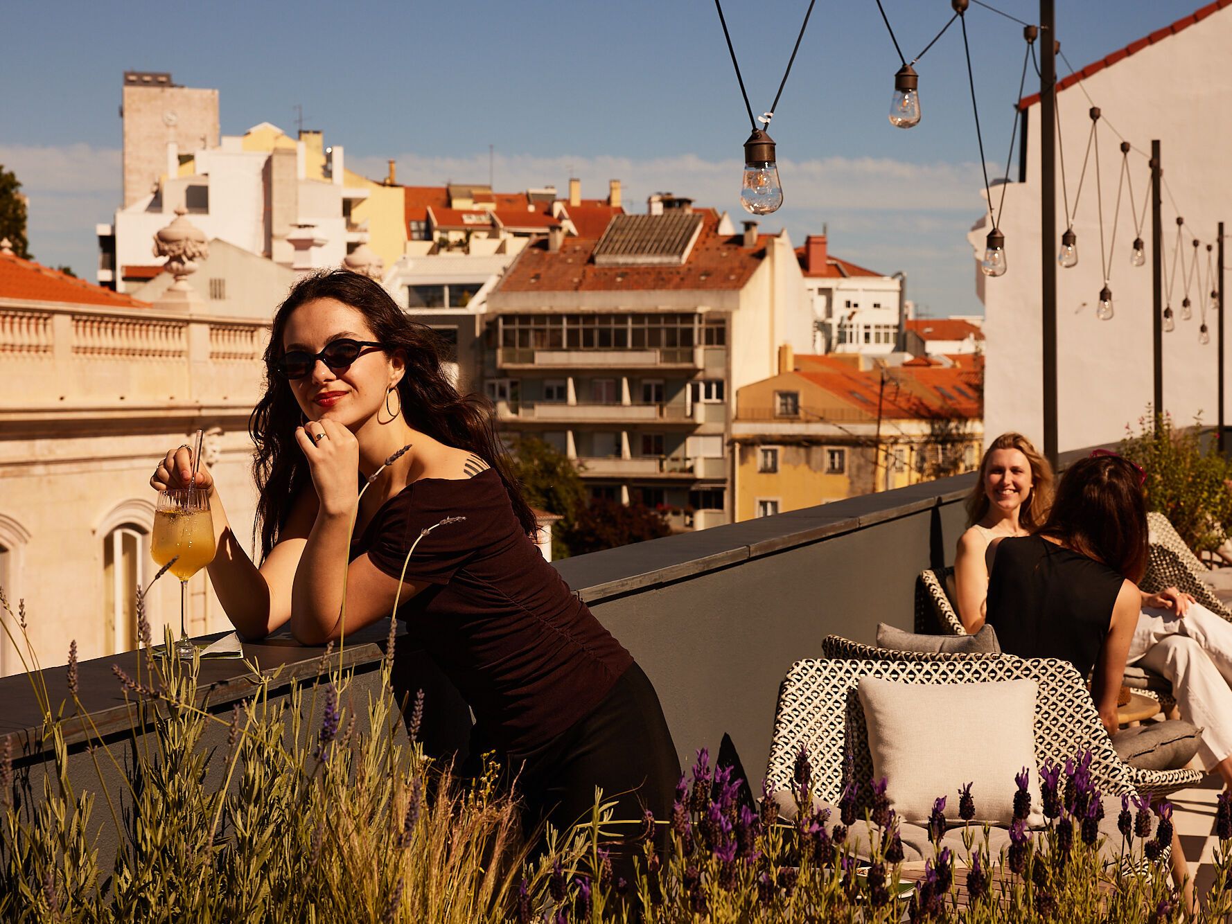 Drei Frauen entspannen auf einer sonnigen Dachterrasse mit Blick auf die Stadt, eine hält ein Getränk, Atmosphäre freundlich und ruhig.