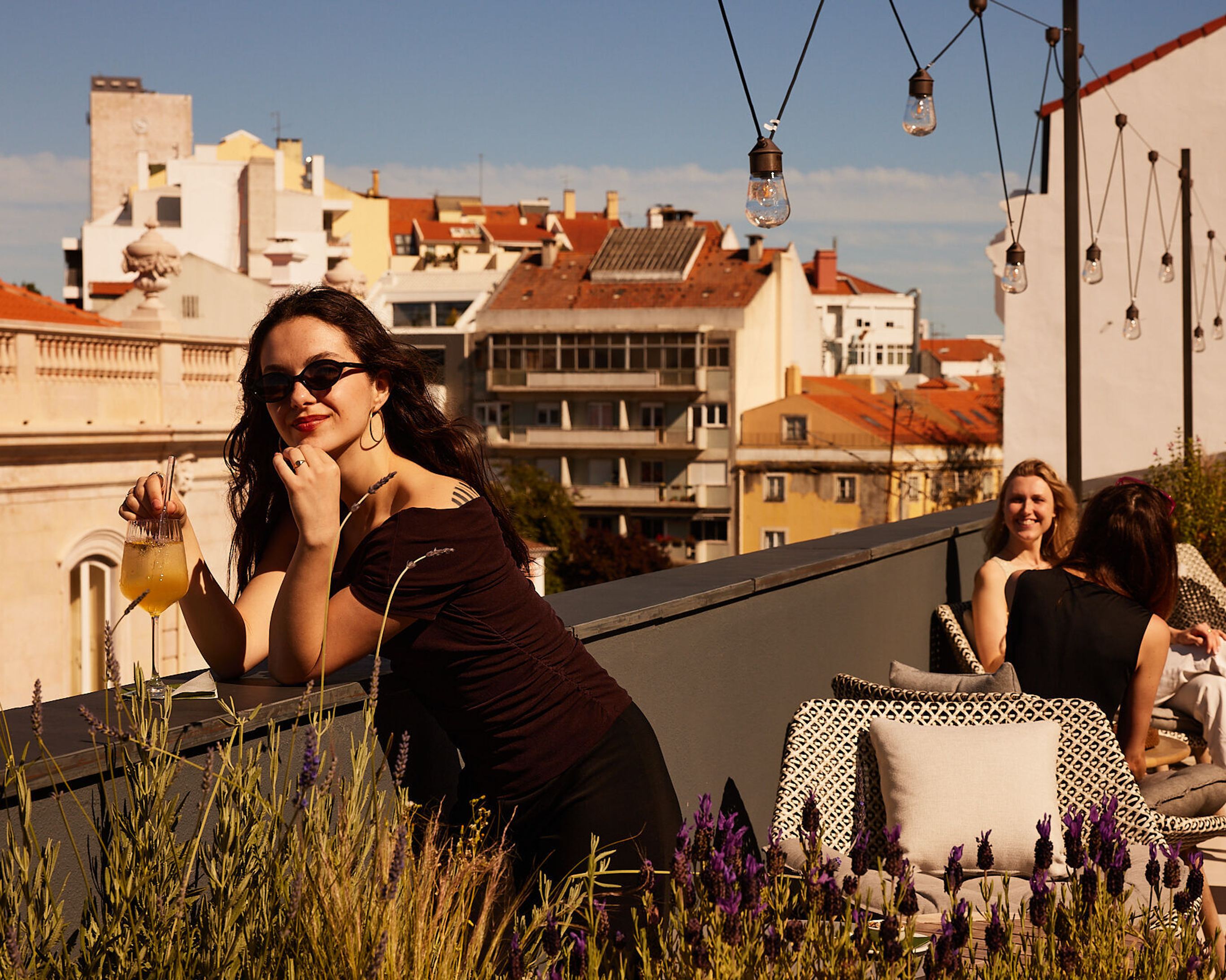 Drei Frauen entspannen auf einer sonnigen Dachterrasse mit Blick auf die Stadt, eine hält ein Getränk, Atmosphäre freundlich und ruhig.