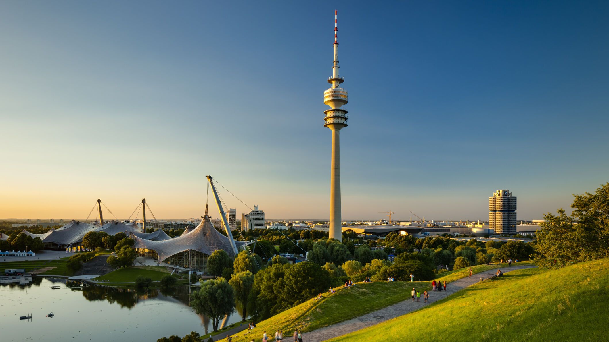 Blick über eine grüne Parklandschaft mit See und Spazierwegen, im Hintergrund der markante Olympiaturm und das Zeltdach des Olympiaparks bei klarem, sonnigem Himmel.