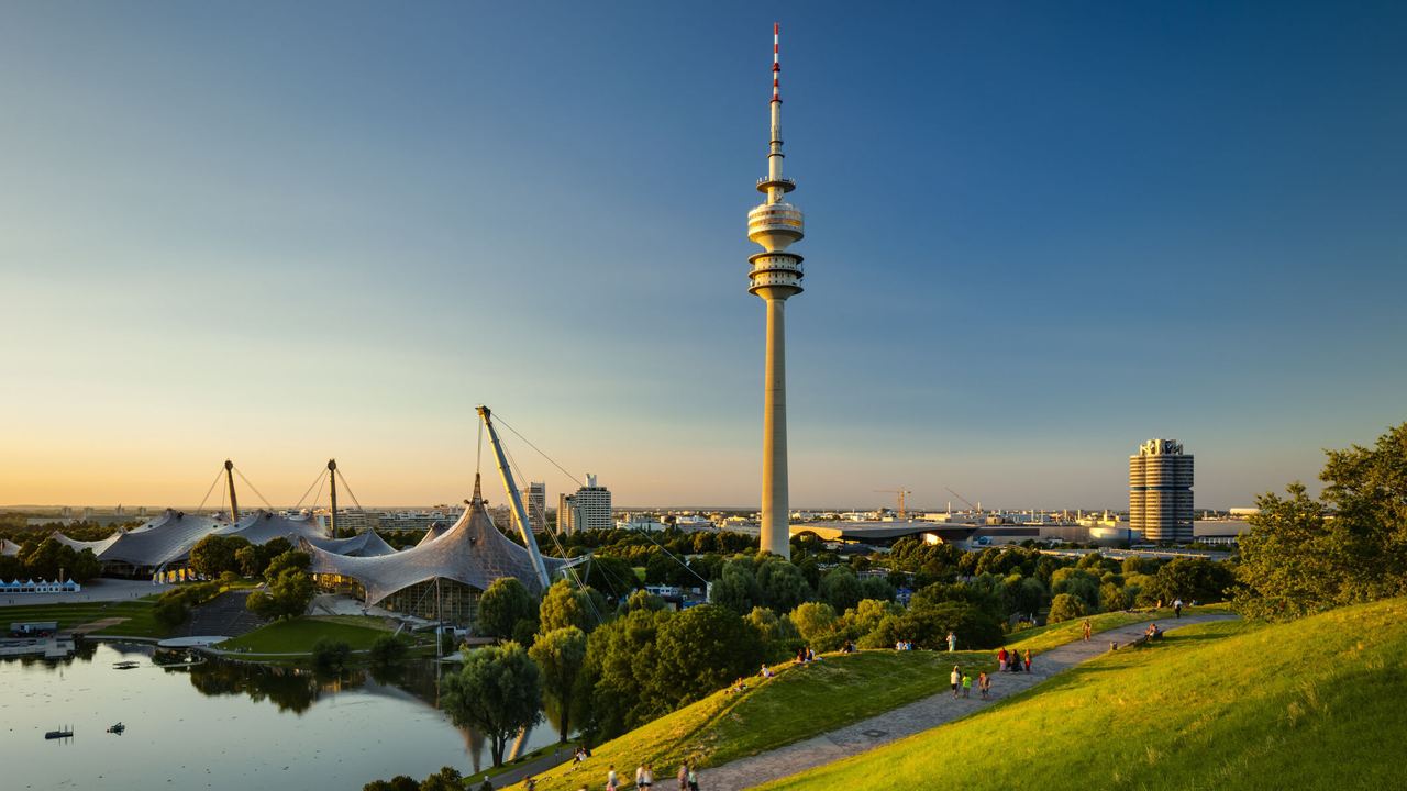 Blick über eine grüne Parklandschaft mit See und Spazierwegen, im Hintergrund der markante Olympiaturm und das Zeltdach des Olympiaparks bei klarem, sonnigem Himmel. Blick über eine grüne Parklandschaft mit See und Spazierwegen, im Hintergrund der markante Olympiaturm und das Zeltdach des Olympiaparks bei klarem, sonnigem Himmel.