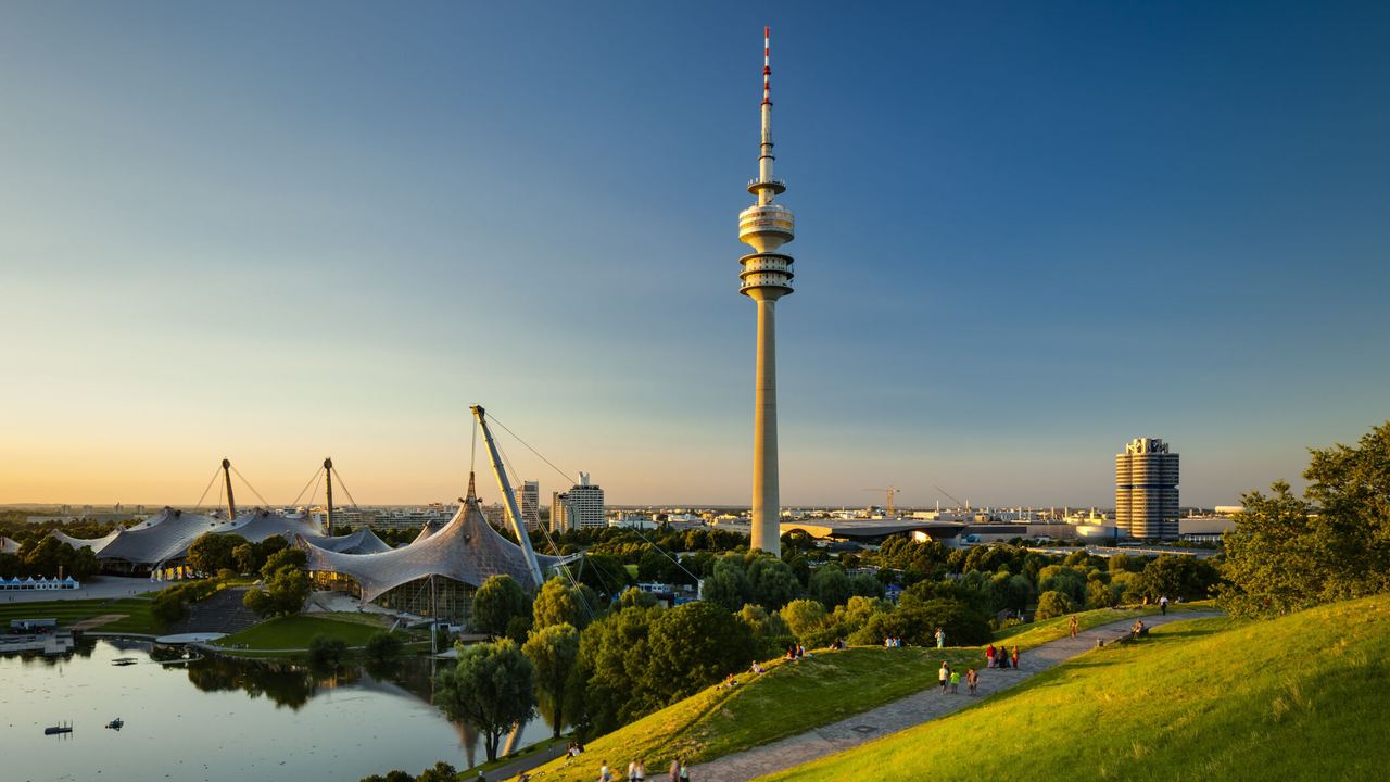 Blick über eine grüne Parklandschaft mit See und Spazierwegen, im Hintergrund der markante Olympiaturm und das Zeltdach des Olympiaparks bei klarem, sonnigem Himmel.