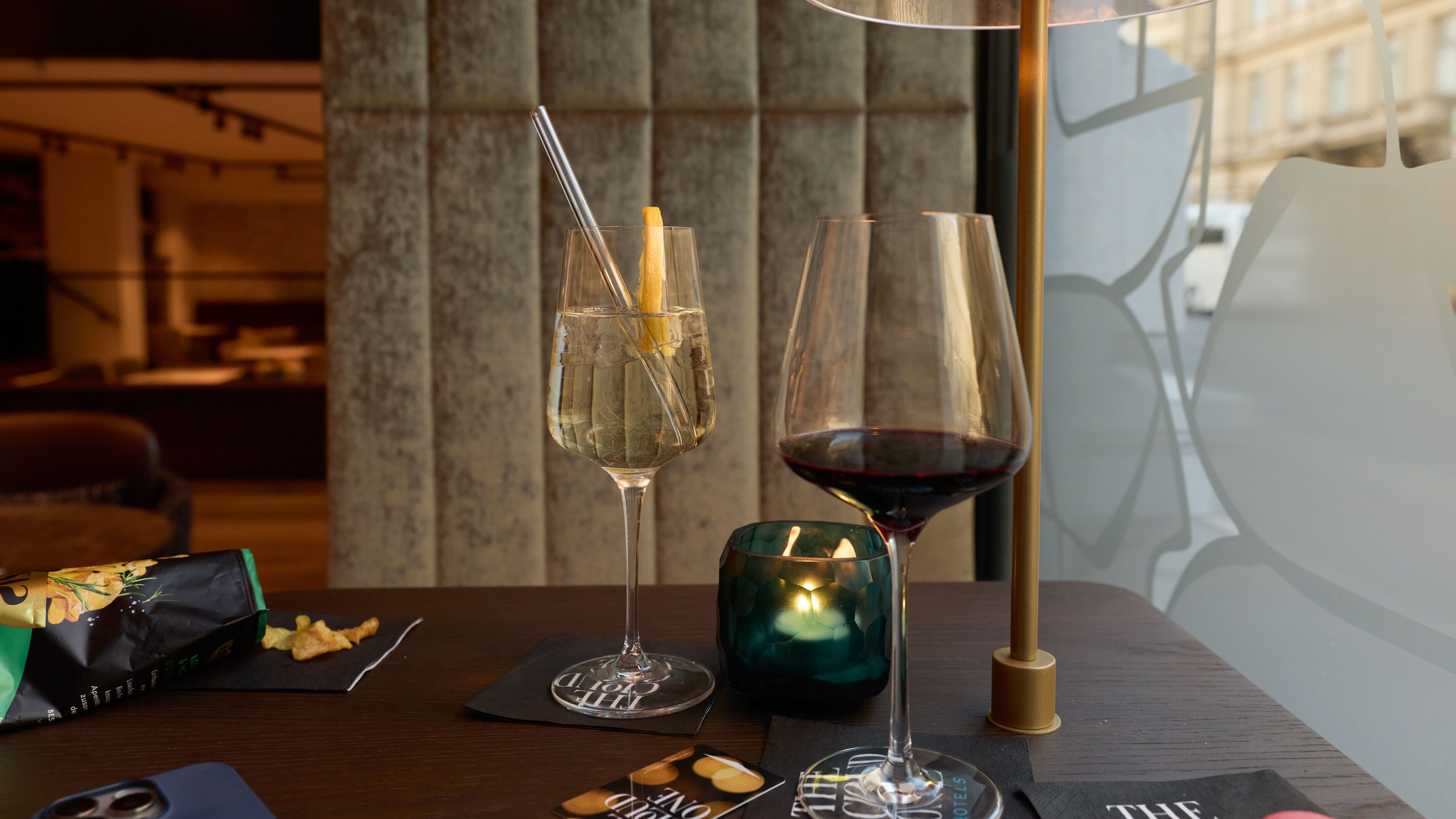 Zwei Getränke, ein Glas Weißwein und ein Glas Rotwein, stehen auf einem Tisch neben einer kleinen Kerze und einer modernen Lampe in einer stilvollen, gemütlichen Bar oder Lounge.