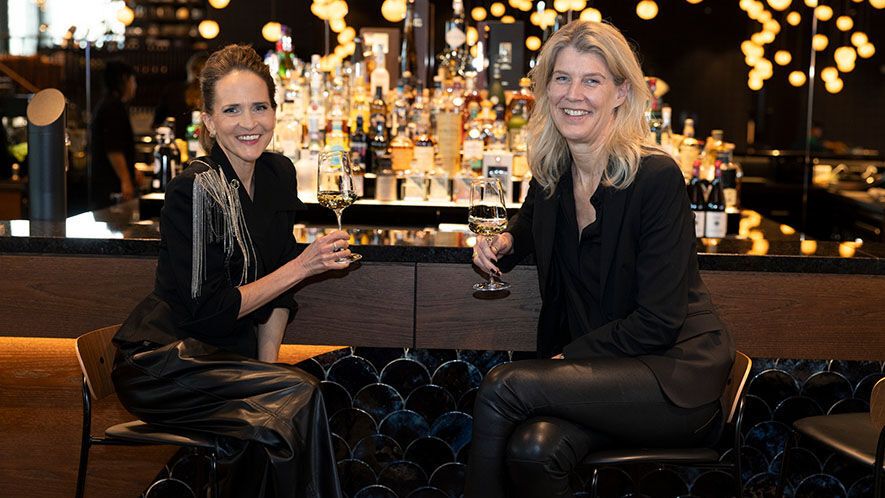 Zwei Frauen sitzen entspannt an einer stilvollen Hotelbar und stoßen mit Weingläsern an. 