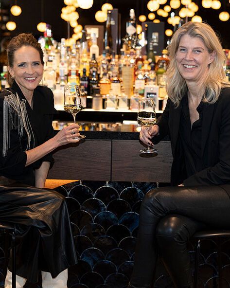 Zwei Frauen sitzen entspannt an einer stilvollen Hotelbar und stoßen mit Weingläsern an. 