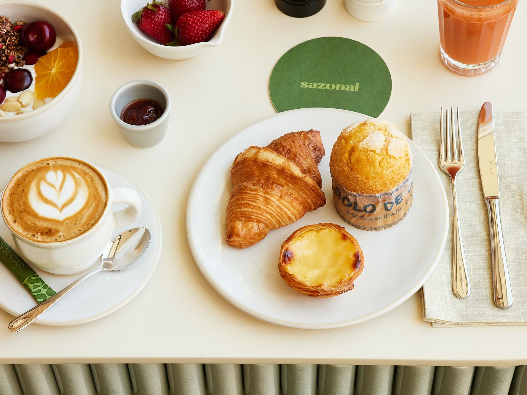 Frühstück mit Cappuccino, Croissant, Pudding-Törtchen, Muffin, frischen Erdbeeren, Joghurt mit Granola und Früchten sowie einem Glas Saft, stilvoll auf hellem Tisch angerichtet.