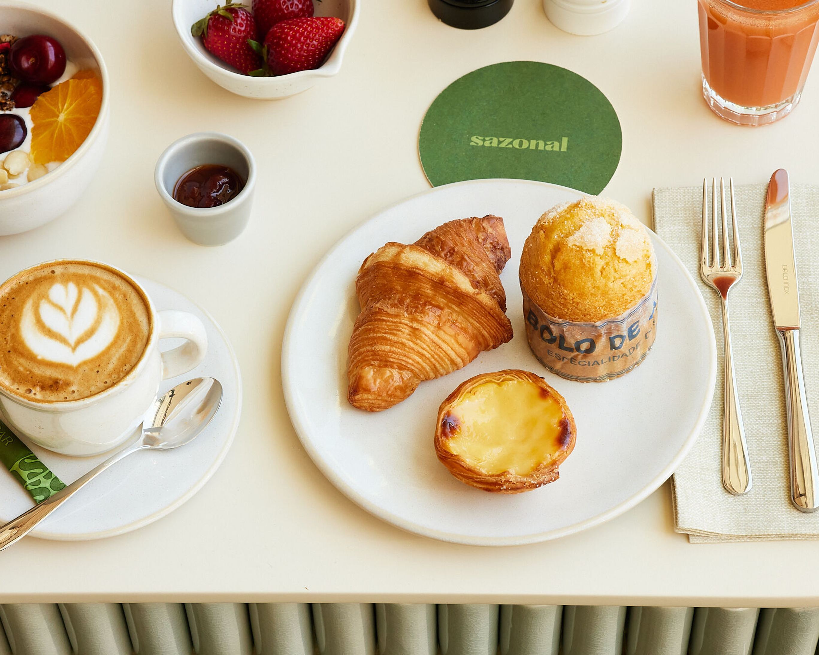 Frühstück mit Cappuccino, Croissant, Pudding-Törtchen, Muffin, frischen Erdbeeren, Joghurt mit Granola und Früchten sowie einem Glas Saft, stilvoll auf hellem Tisch angerichtet.