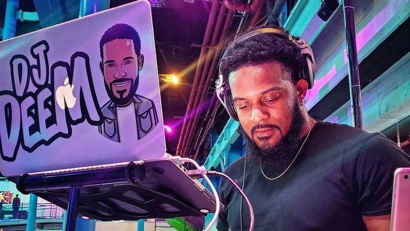 Ein DJ mit Kopfhörern mischt fokussiert Musik an einem modernen DJ-Set in einem lebhaften Club oder Veranstaltungsraum. Sein Laptop zeigt eine Grafik mit dem Namen „DJ DEEM“. Die Szene strahlt energiegeladene Stimmung und musikalischen Flair aus.