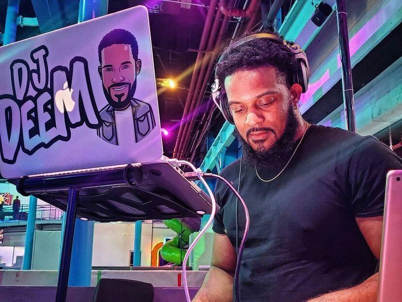 Ein DJ mit Kopfhörern mischt fokussiert Musik an einem modernen DJ-Set in einem lebhaften Club oder Veranstaltungsraum. Sein Laptop zeigt eine Grafik mit dem Namen „DJ DEEM“. Die Szene strahlt energiegeladene Stimmung und musikalischen Flair aus.