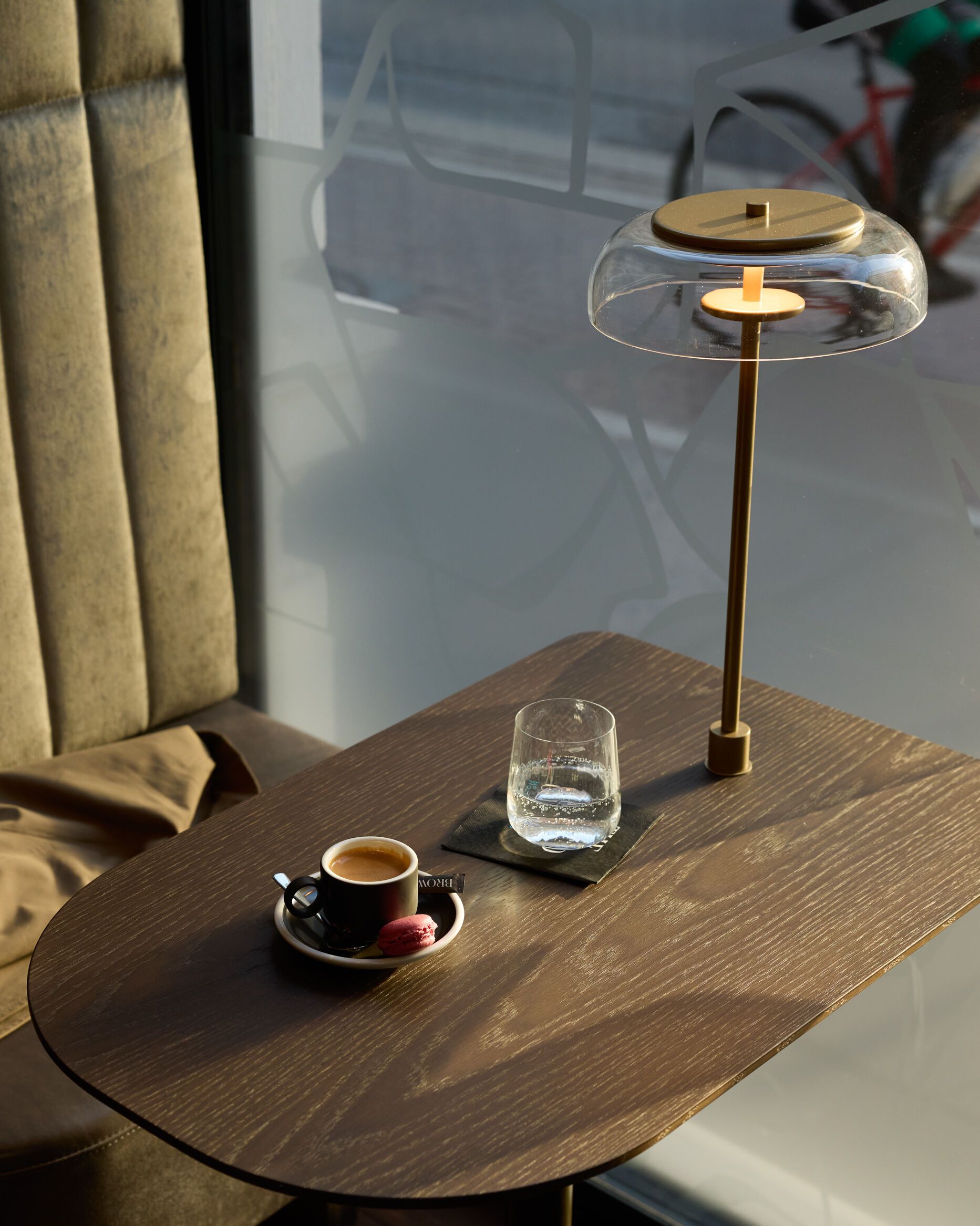 Gemütliche Sitzecke mit samtbezogenem Polstersessel, einem kleinen Holztisch mit Espresso, Glas Wasser und moderner Tischlampe, im Hintergrund Tageslicht durch ein Fenster und Blick auf eine Straße.