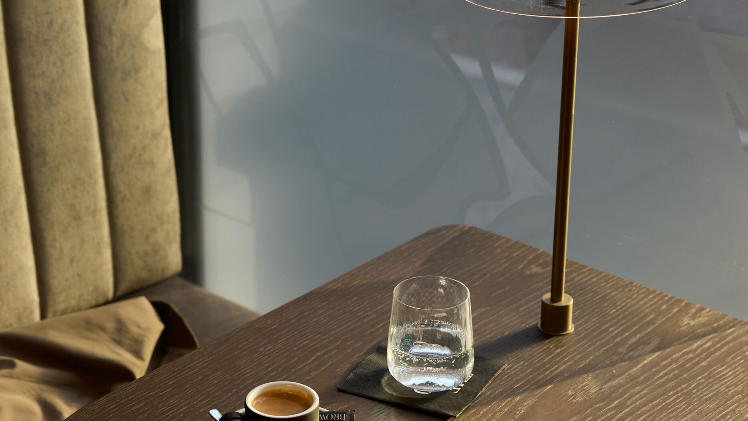 Gemütliche Sitzecke mit samtbezogenem Polstersessel, einem kleinen Holztisch mit Espresso, Glas Wasser und moderner Tischlampe, im Hintergrund Tageslicht durch ein Fenster und Blick auf eine Straße. Gemütliche Sitzecke mit samtbezogenem Polstersessel, einem kleinen Holztisch mit Espresso, Glas Wasser und moderner Tischlampe, im Hintergrund Tageslicht durch ein Fenster und Blick auf eine Straße.