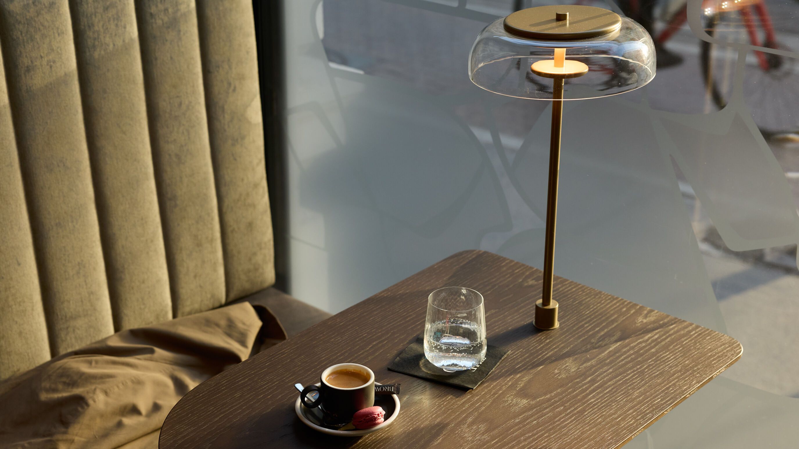 Gemütliche Sitzecke mit samtbezogenem Polstersessel, einem kleinen Holztisch mit Espresso, Glas Wasser und moderner Tischlampe, im Hintergrund Tageslicht durch ein Fenster und Blick auf eine Straße.