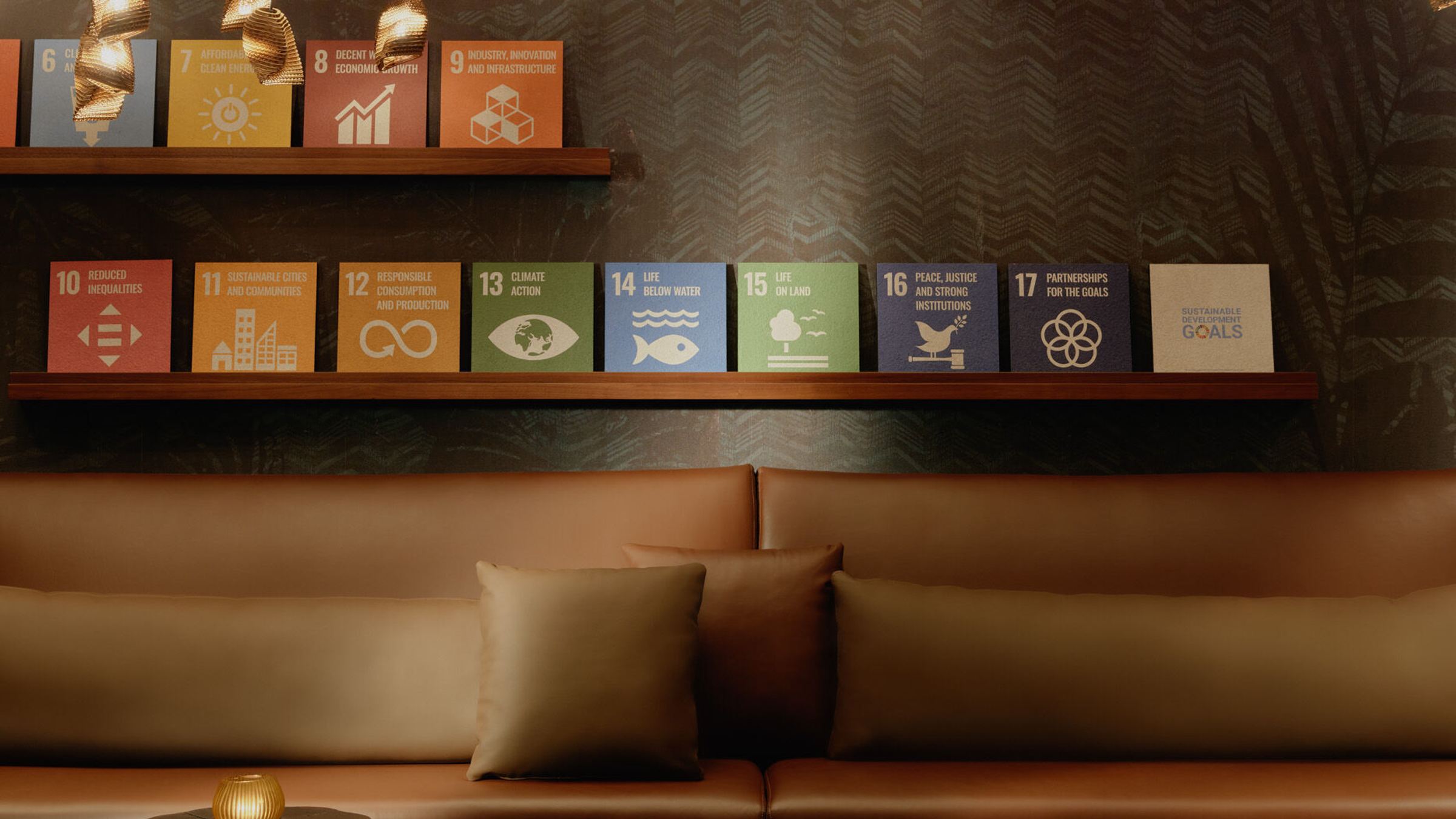 Gemütliche Lounge mit braunem Ledersofa, dekorativen Kissen und dreifach unterschiedlich hohen Holztischen. Über der Sitzgruppe hängen zahlreiche warme, goldene Hängeleuchten, darunter bunte Poster mit Symbolen für nachhaltige Entwicklungsziele an einer dunklen Wand.