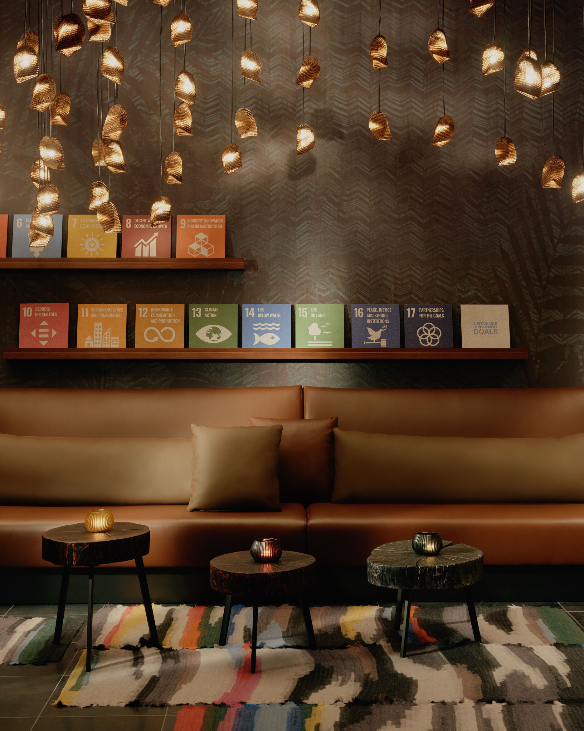 Gemütliche Lounge mit braunem Ledersofa, dekorativen Kissen und dreifach unterschiedlich hohen Holztischen. Über der Sitzgruppe hängen zahlreiche warme, goldene Hängeleuchten, darunter bunte Poster mit Symbolen für nachhaltige Entwicklungsziele an einer dunklen Wand.
