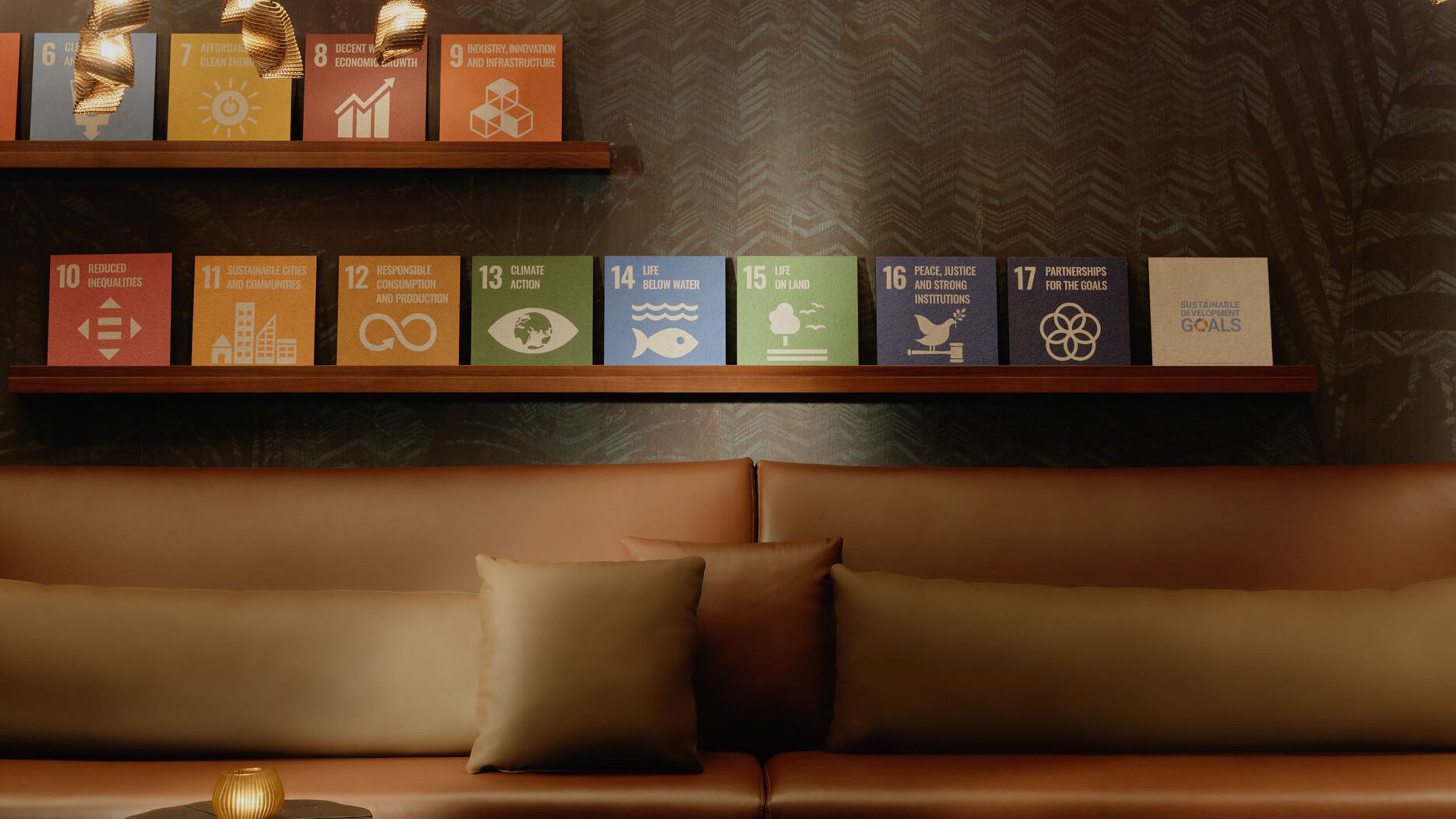 Gemütliche Lounge mit braunem Ledersofa, dekorativen Kissen und dreifach unterschiedlich hohen Holztischen. Über der Sitzgruppe hängen zahlreiche warme, goldene Hängeleuchten, darunter bunte Poster mit Symbolen für nachhaltige Entwicklungsziele an einer dunklen Wand. Gemütliche Lounge mit braunem Ledersofa, dekorativen Kissen und dreifach unterschiedlich hohen Holztischen. Über der Sitzgruppe hängen zahlreiche warme, goldene Hängeleuchten, darunter bunte Poster mit Symbolen für nachhaltige Entwicklungsziele an einer dunklen Wand.