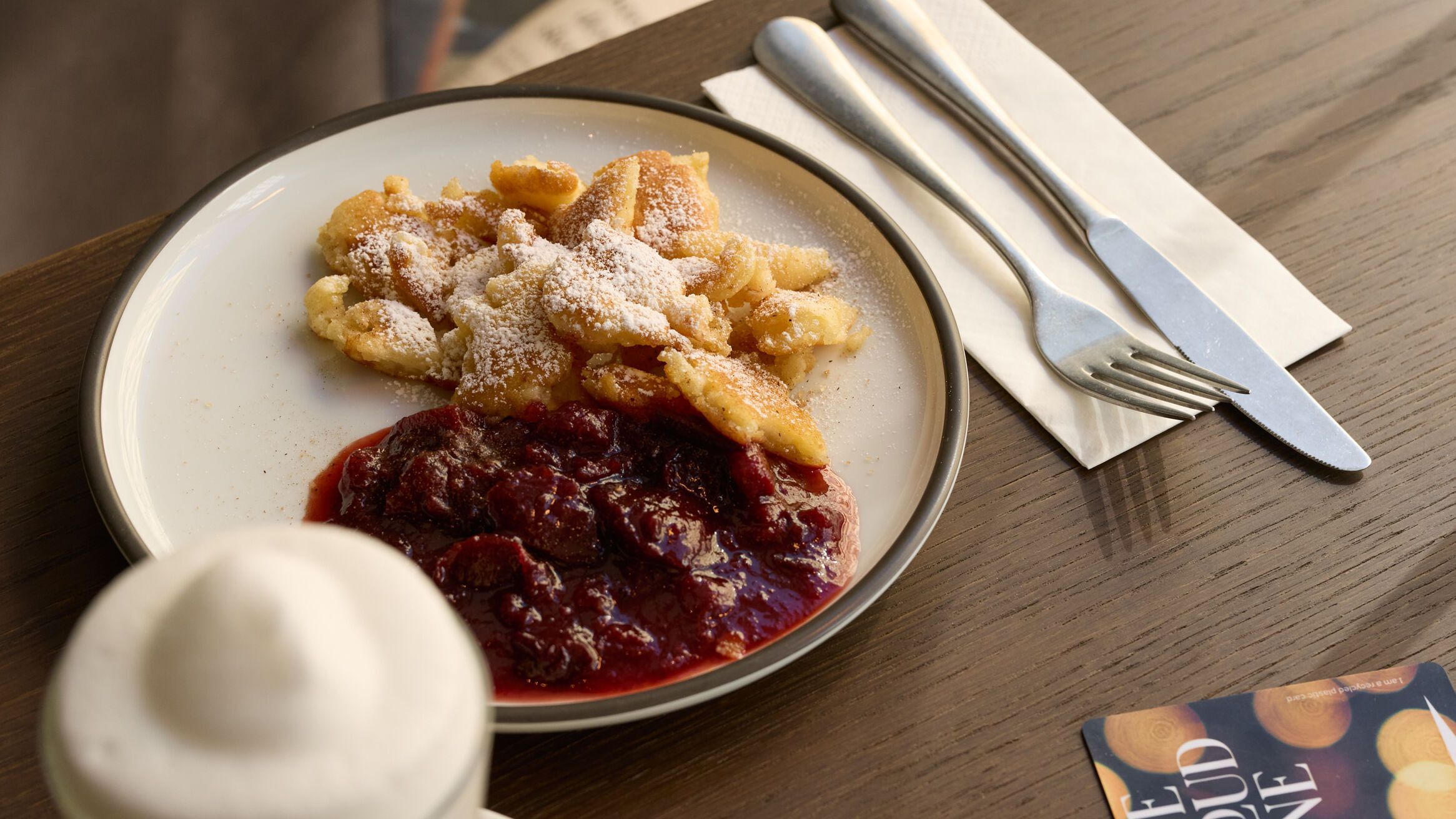 Ein Teller mit Kaiserschmarrn und Zwetschgenröster auf einem Holztisch, daneben ein Cappuccino mit Milchschaum und Besteck auf einer Serviette, gemütliche Sitzgelegenheiten im Hintergrund.