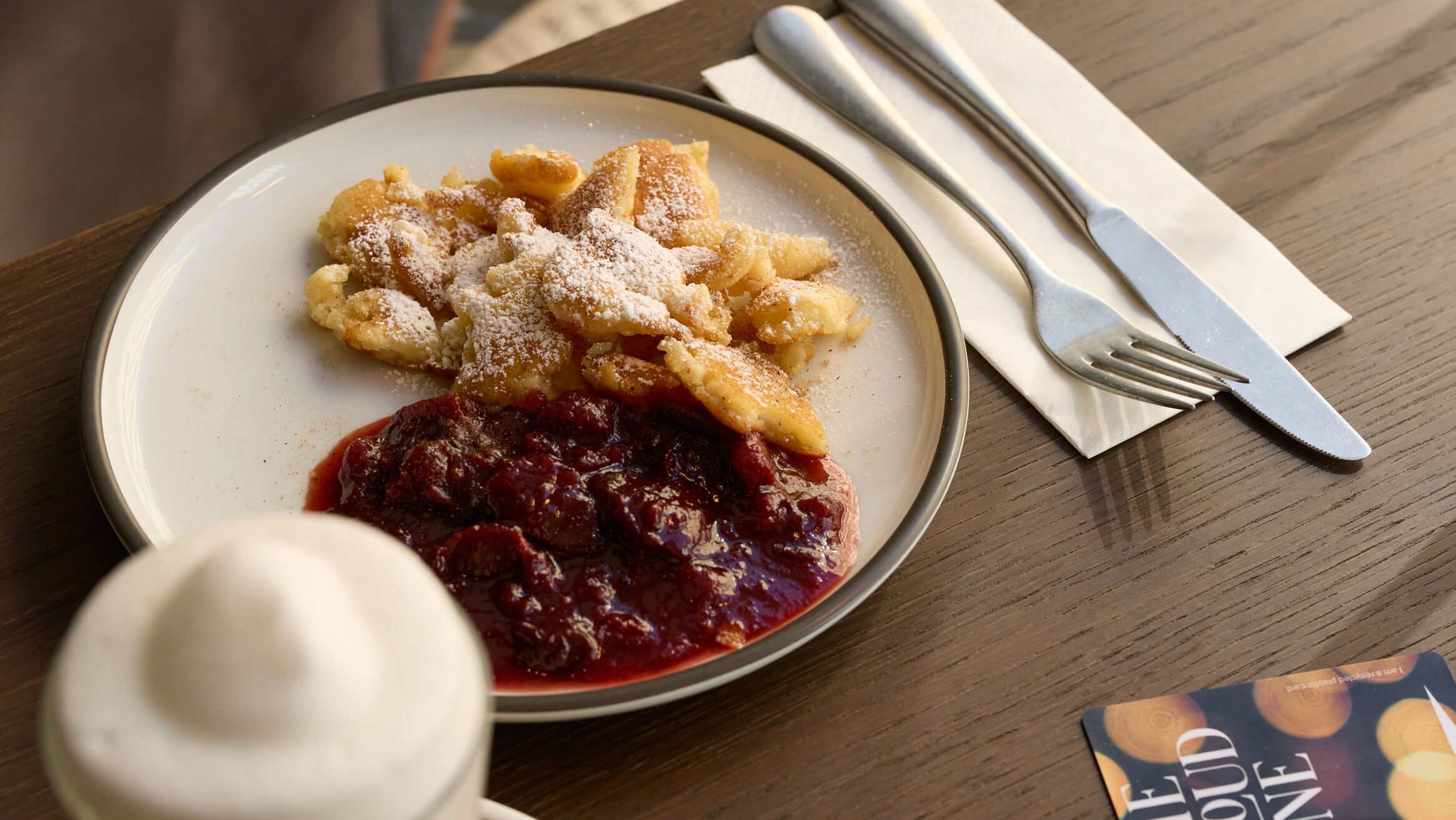 Ein Teller mit Kaiserschmarrn und Zwetschgenröster auf einem Holztisch, daneben ein Cappuccino mit Milchschaum und Besteck auf einer Serviette, gemütliche Sitzgelegenheiten im Hintergrund.