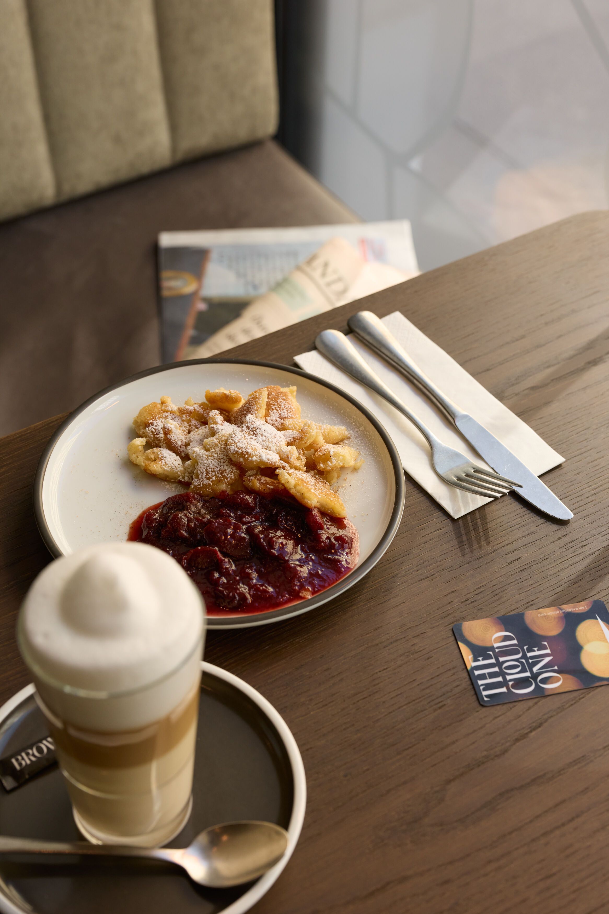 Ein Teller mit Kaiserschmarrn und Zwetschgenröster auf einem Holztisch, daneben ein Cappuccino mit Milchschaum und Besteck auf einer Serviette, gemütliche Sitzgelegenheiten im Hintergrund.