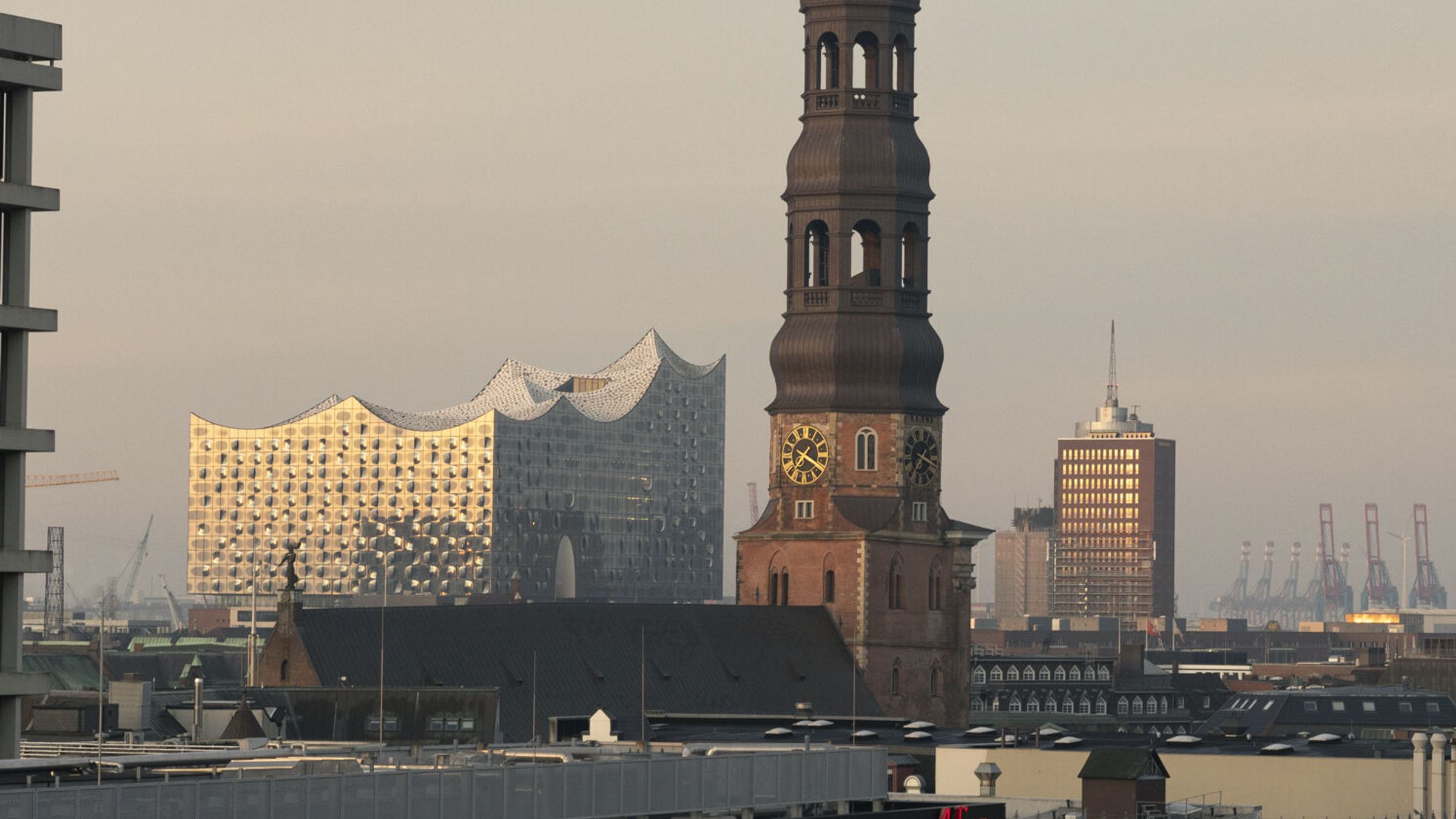 Blick über die Dächer Hamburgs mit der markanten Elbphilharmonie, dem Turm der St. Katharinenkirche und Hafenkränen im Hintergrund, bei ruhigem, goldenem Abendlicht. Blick über die Dächer Hamburgs mit der markanten Elbphilharmonie, dem Turm der St. Katharinenkirche und Hafenkränen im Hintergrund, bei ruhigem, goldenem Abendlicht.