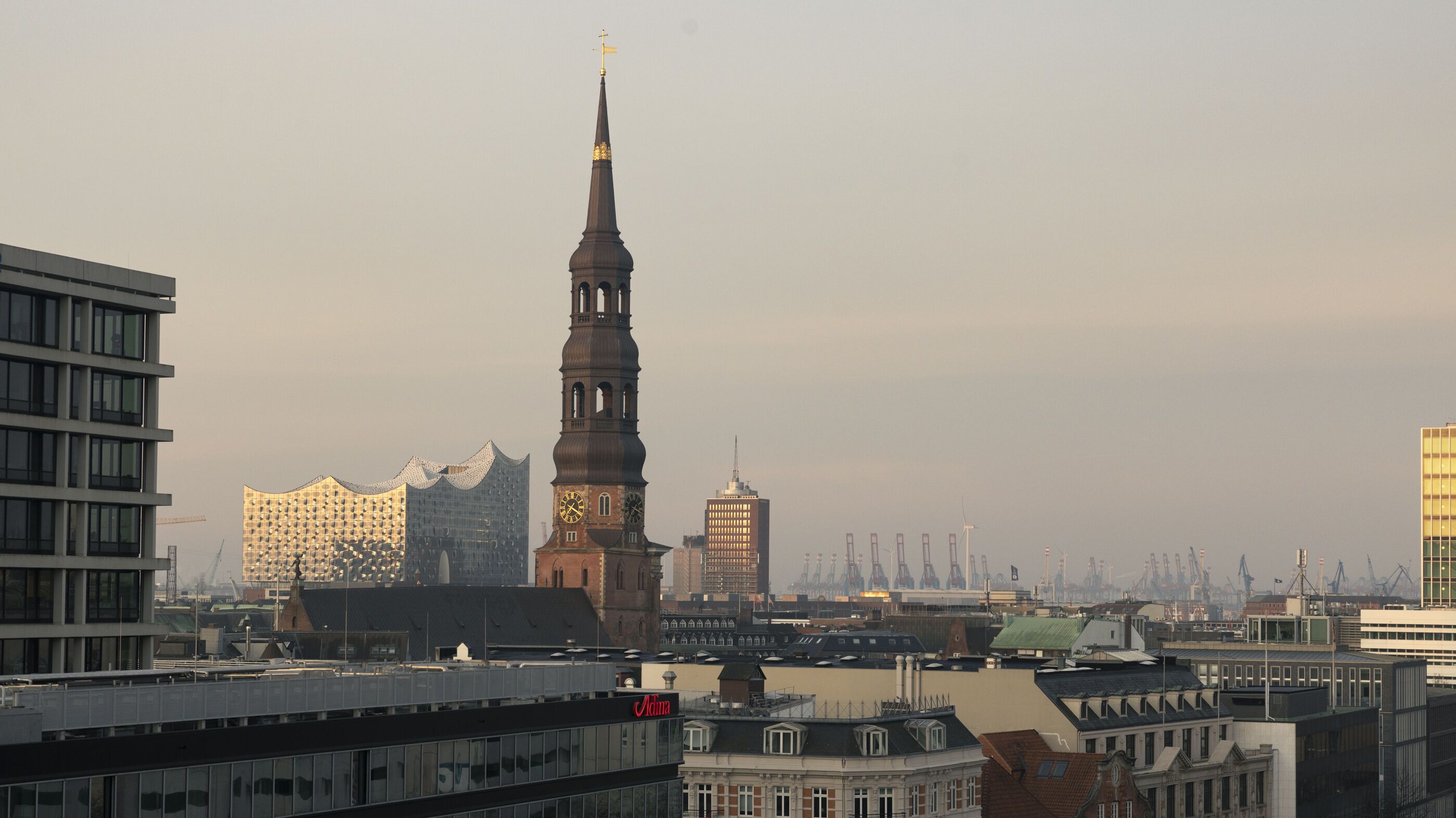 Blick über die Dächer Hamburgs mit der markanten Elbphilharmonie, dem Turm der St. Katharinenkirche und Hafenkränen im Hintergrund, bei ruhigem, goldenem Abendlicht.