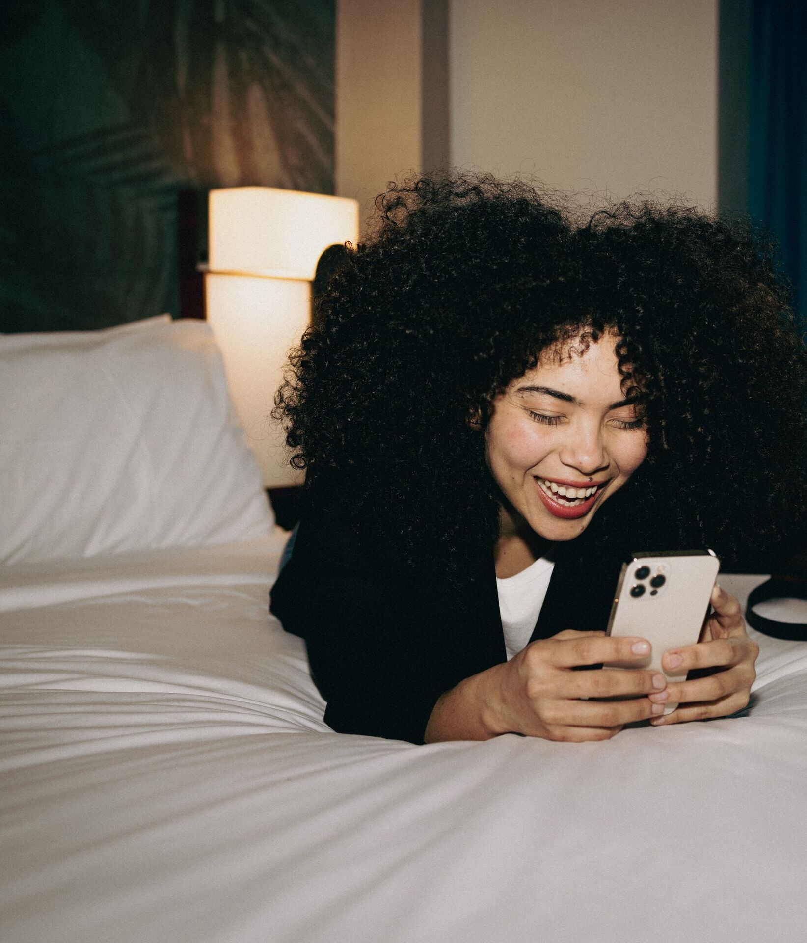 Frau liegt entspannt auf einem Hotelbett und lächelt beim Blick auf ihr Smartphone, gemütliche Atmosphäre im Zimmer mit warmem Licht.