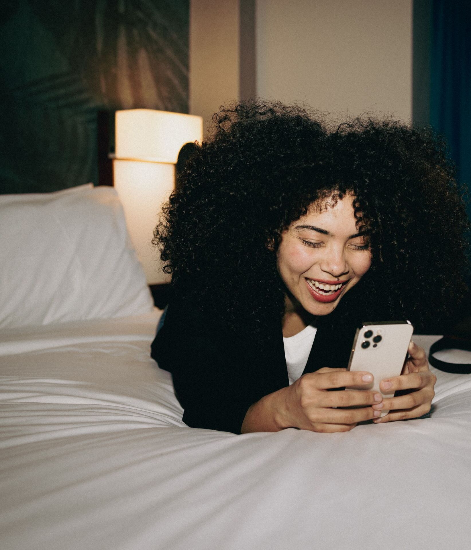 Frau liegt entspannt auf einem Hotelbett und lächelt beim Blick auf ihr Smartphone, gemütliche Atmosphäre im Zimmer mit warmem Licht.