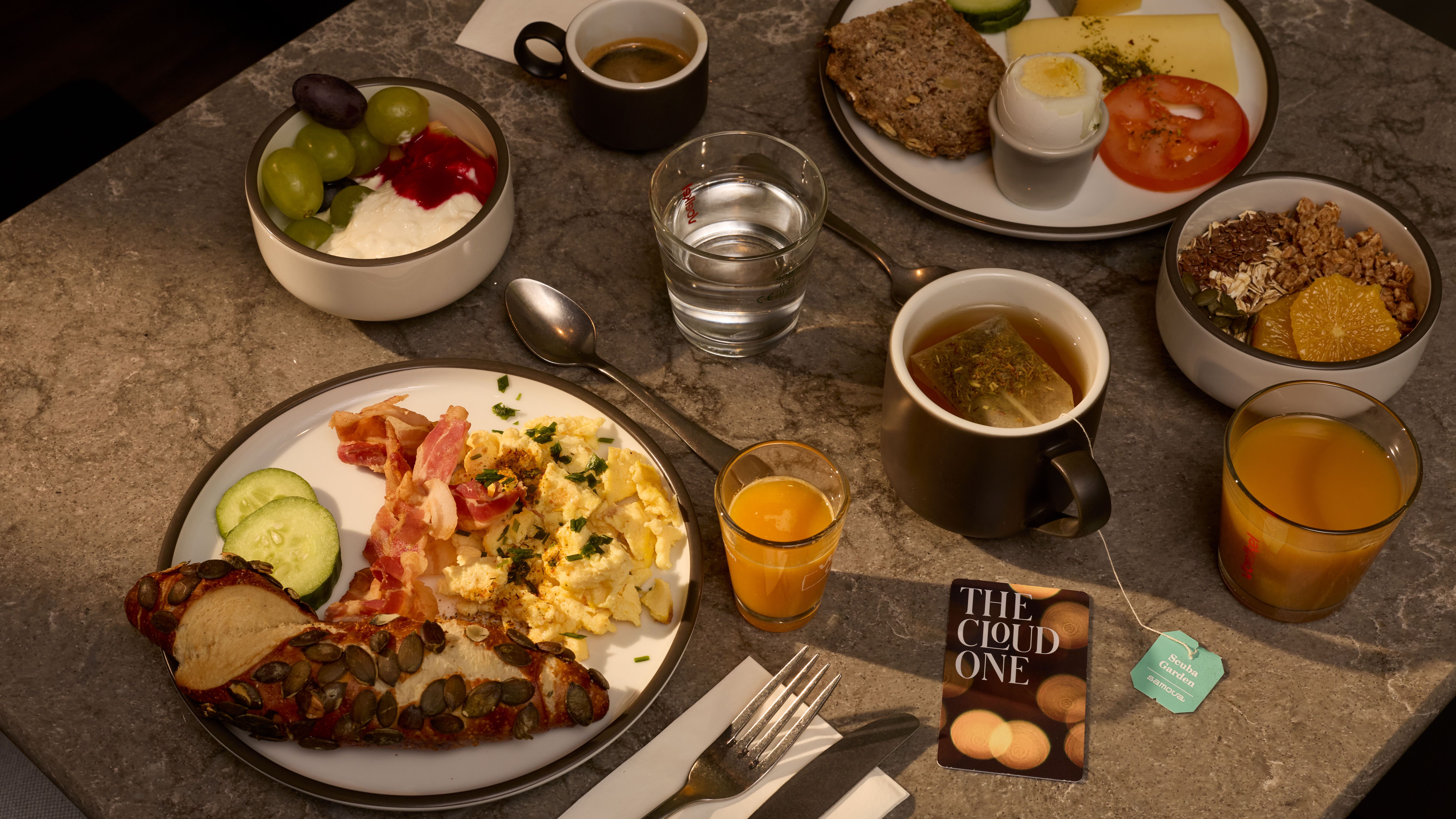 Frühstückstisch mit Rührei, Speck, Gurkenscheiben, Kürbiskernbrot, Joghurt mit Trauben, Müsli mit Honig, gekochtem Ei, Tomate, Käse, Kaffee, Tee, Orangensaft und Wasser.