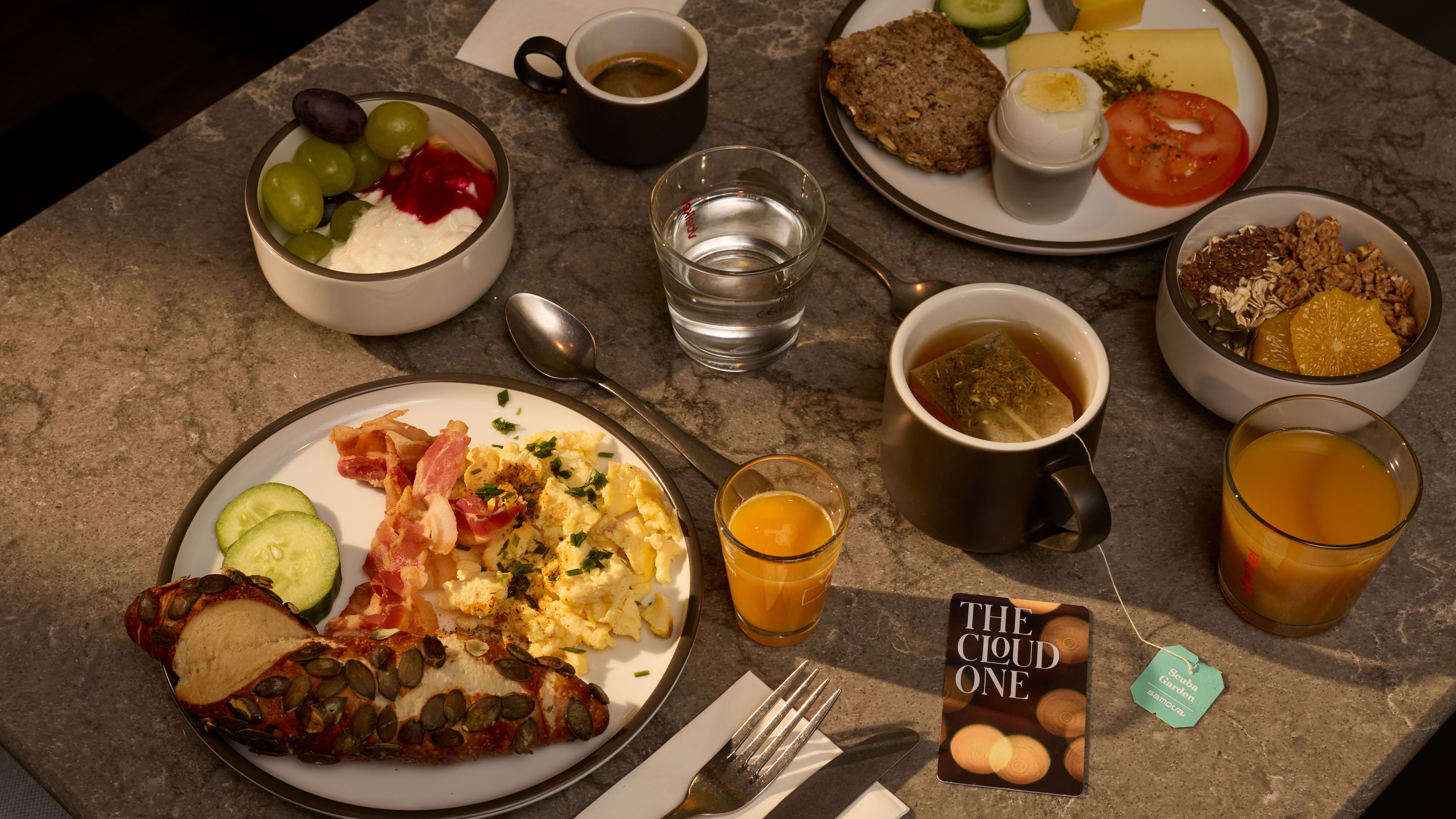 Frühstückstisch mit Rührei, Speck, Gurkenscheiben, Kürbiskernbrot, Joghurt mit Trauben, Müsli mit Honig, gekochtem Ei, Tomate, Käse, Kaffee, Tee, Orangensaft und Wasser.