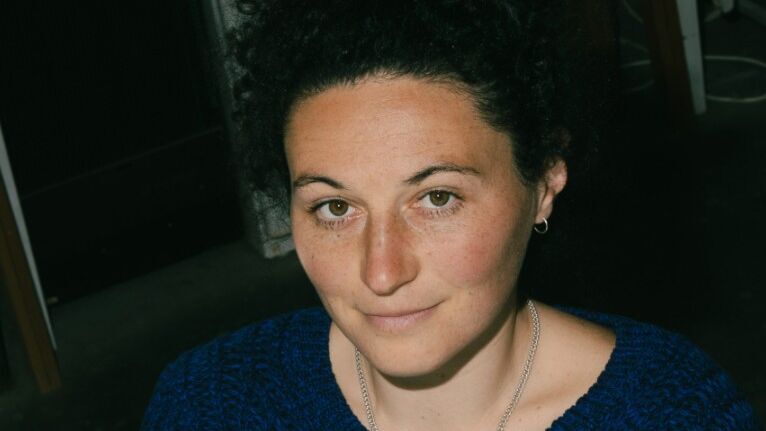 Porträt einer Frau mit lockigem, dunklem Haar, die lächelnd und mit verschränkten Armen in einem gemütlichen blauen Pullover sitzt.