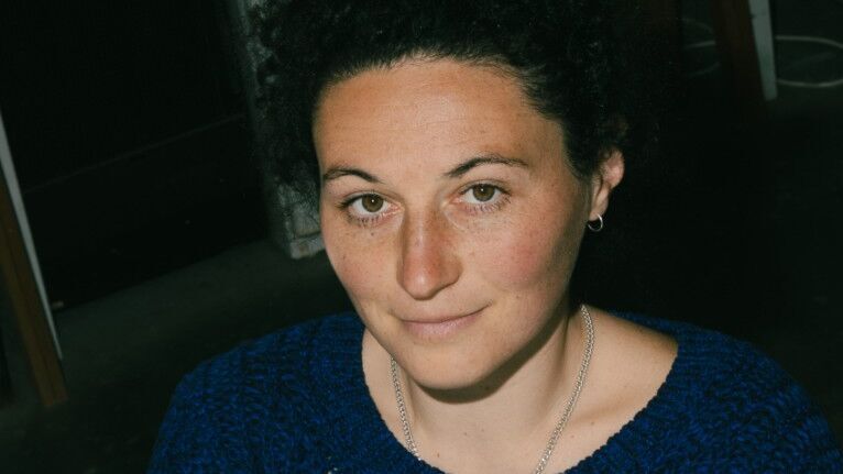 Porträt einer Frau mit lockigem, dunklem Haar, die lächelnd und mit verschränkten Armen in einem gemütlichen blauen Pullover sitzt.