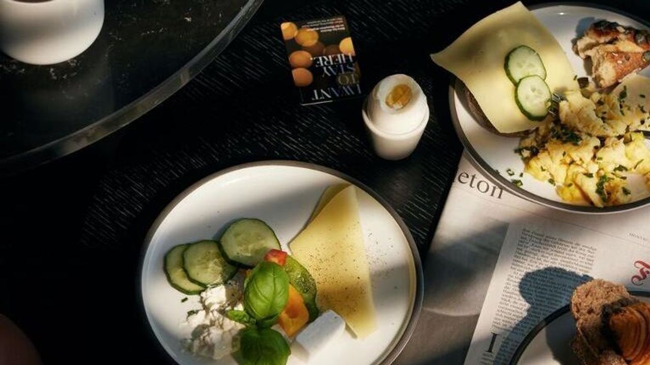 Frühstück mit Croissants, Käse, Gurkenscheiben, Rührei und Cappuccino, angerichtet auf weißen Tellern und serviert in natürlichem Licht.