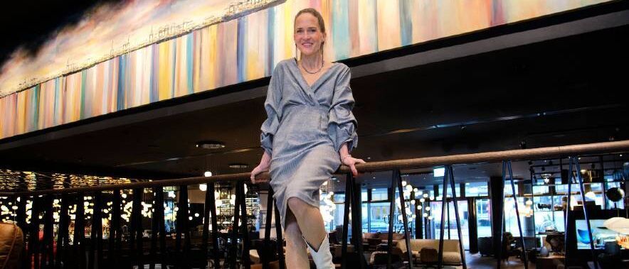 Eine Frau in elegantem, silbernem Kleid und weißen Stiefeln sitzt entspannt auf einem Geländer in einem stilvoll beleuchteten Hotel- oder Restaurantbereich.