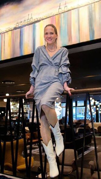 Eine Frau in elegantem, silbernem Kleid und weißen Stiefeln sitzt entspannt auf einem Geländer in einem stilvoll beleuchteten Hotel- oder Restaurantbereich.