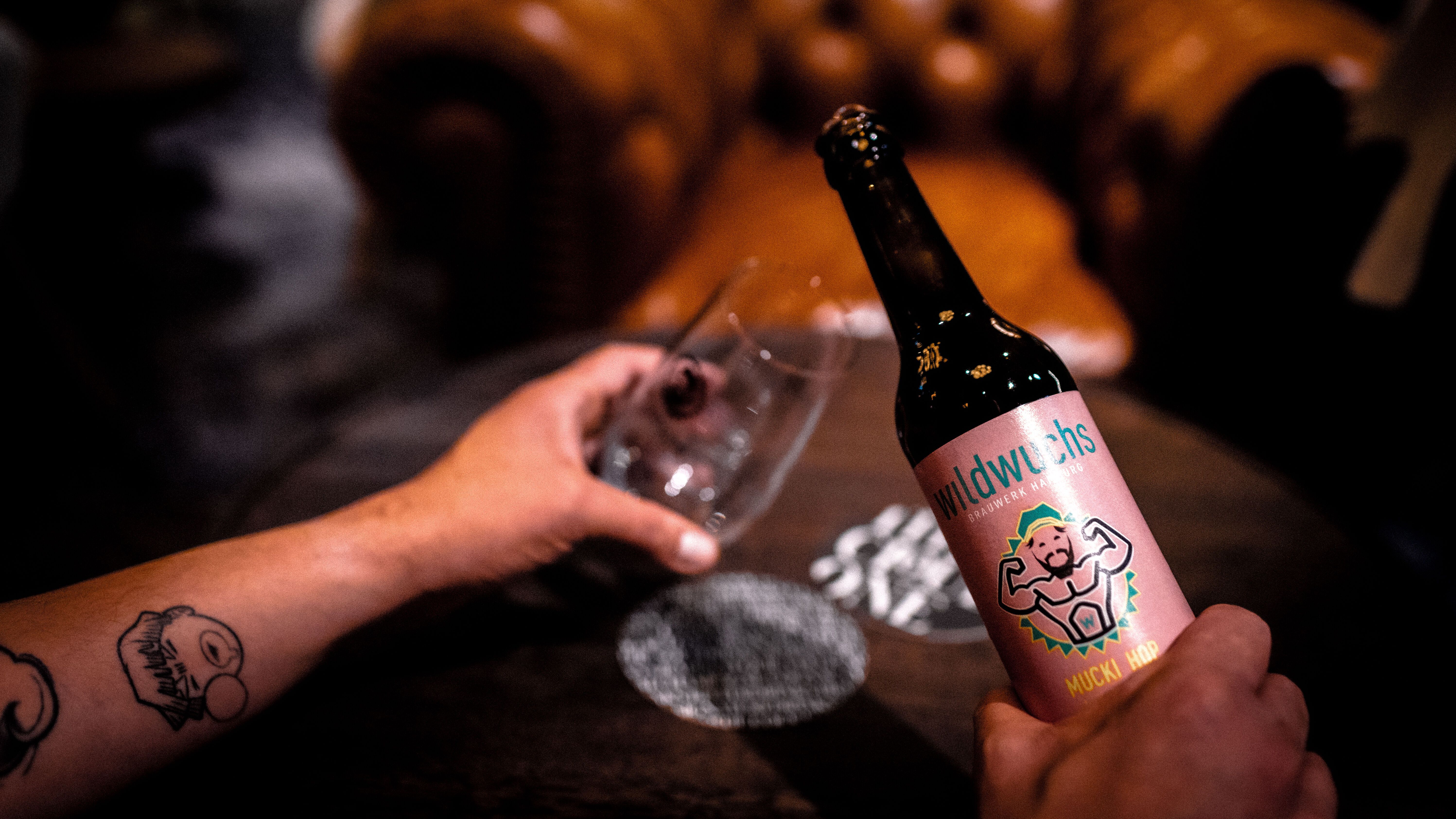 Person hält eine Bierflasche mit farbenfrohem Etikett und ein Glas, im Hintergrund ein gemütliches, braunes Ledersofa, Atmosphäre wirkt entspannt und einladend.