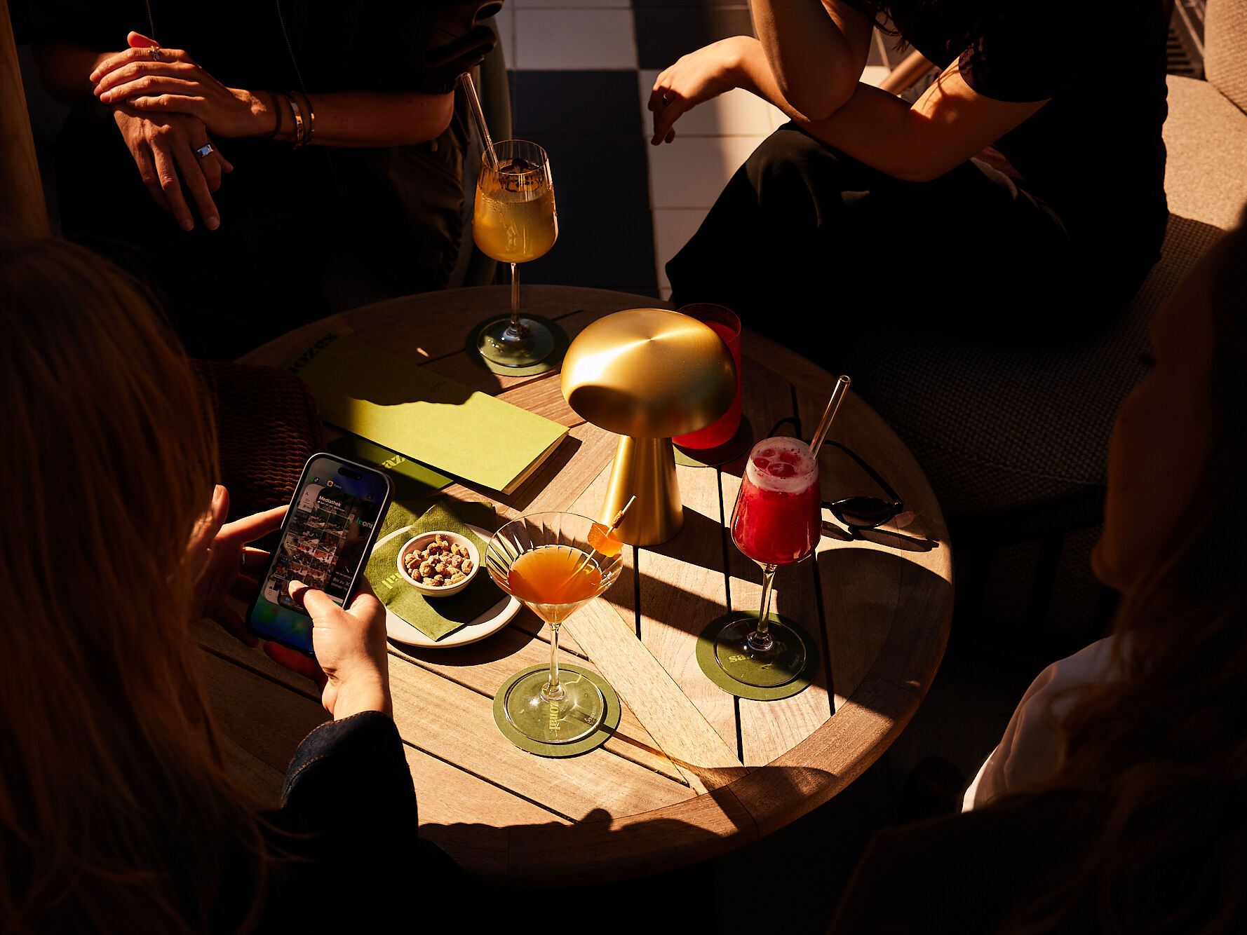 Drei Personen sitzen an einem runden Holztisch mit bunten Cocktails und Snacks, die entspannte Atmosphäre lädt zum gemeinsamen Genießen und Verweilen ein.