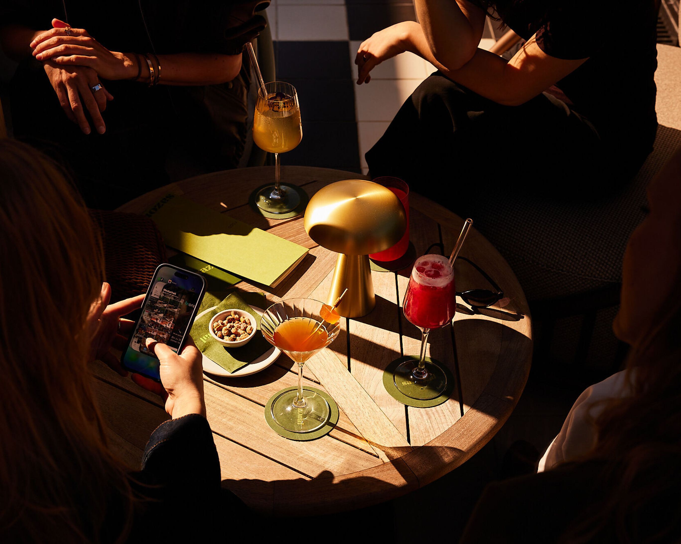 Drei Personen sitzen an einem runden Holztisch mit bunten Cocktails und Snacks, die entspannte Atmosphäre lädt zum gemeinsamen Genießen und Verweilen ein.