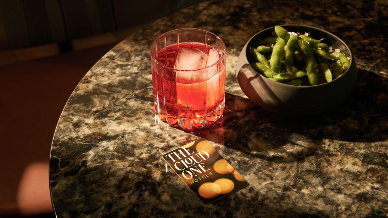 Ein Glas roter Cocktail mit Eiswürfeln und eine Schale grüner Edamame stehen auf einem dunkel marmorierten Tisch neben einer Hotelkarte von "The Cloud One Hotels".