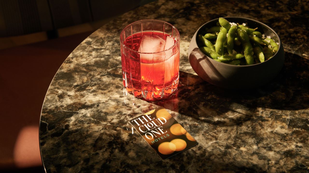 Ein Glas roter Cocktail mit Eiswürfeln und eine Schale grüner Edamame stehen auf einem dunkel marmorierten Tisch neben einer Hotelkarte von "The Cloud One Hotels". Ein Glas roter Cocktail mit Eiswürfeln und eine Schale grüner Edamame stehen auf einem dunkel marmorierten Tisch neben einer Hotelkarte von "The Cloud One Hotels".