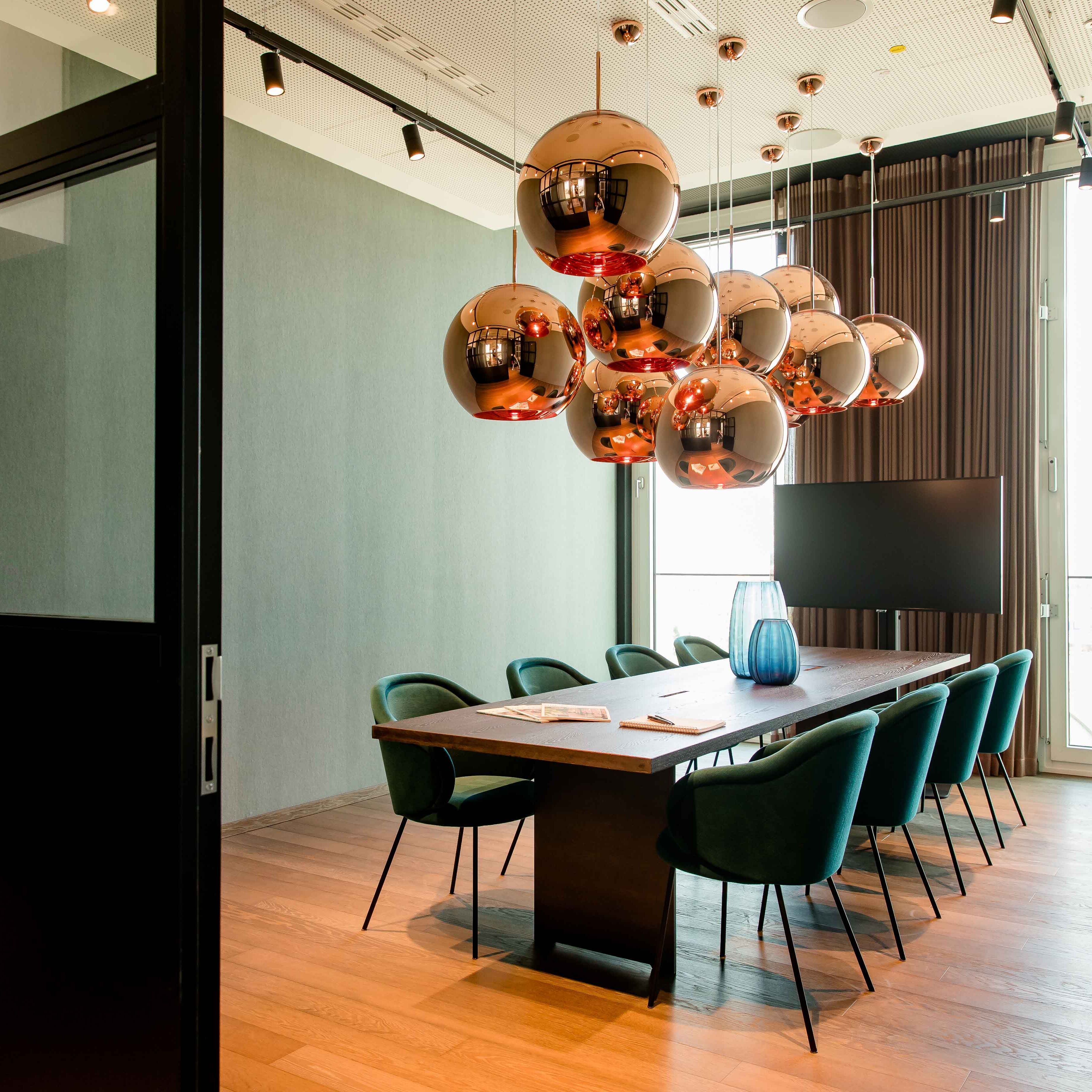 Motel One Konferenzraum mit langem Holztisch, acht grünen Stühlen und kupferfarbenen Kugelpendelleuchten, hell durch großflächige Fenster mit Tageslicht erleuchtet.